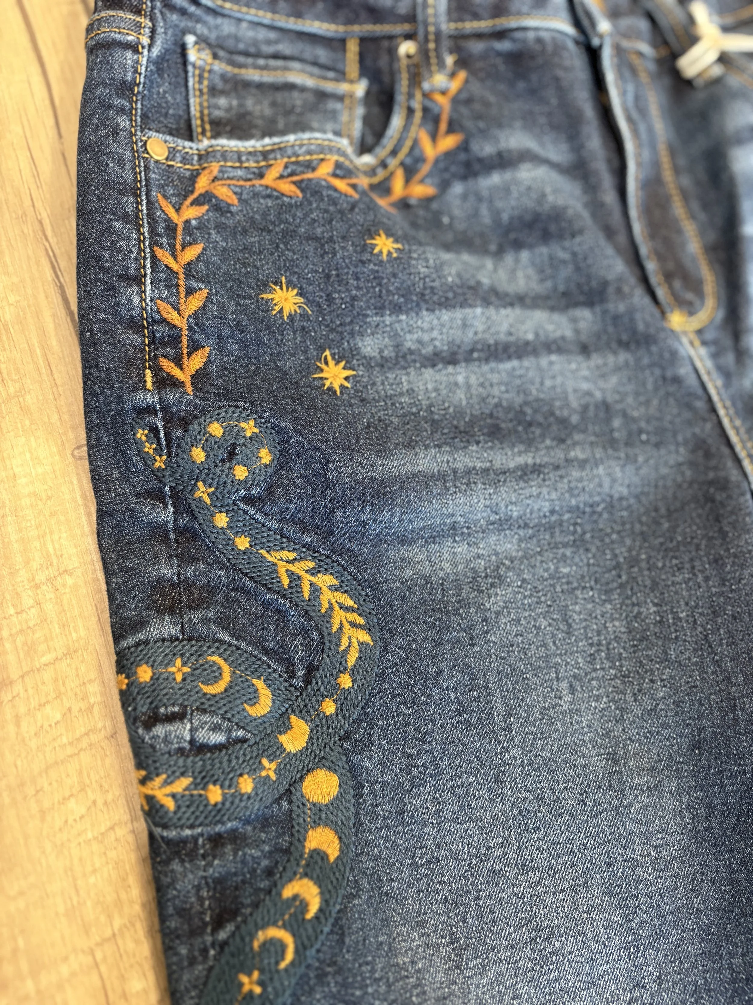 Close-up of blue denim jeans with yellow embroidery on the pocket and side seam, showing a floral design with leaves and small flowers.