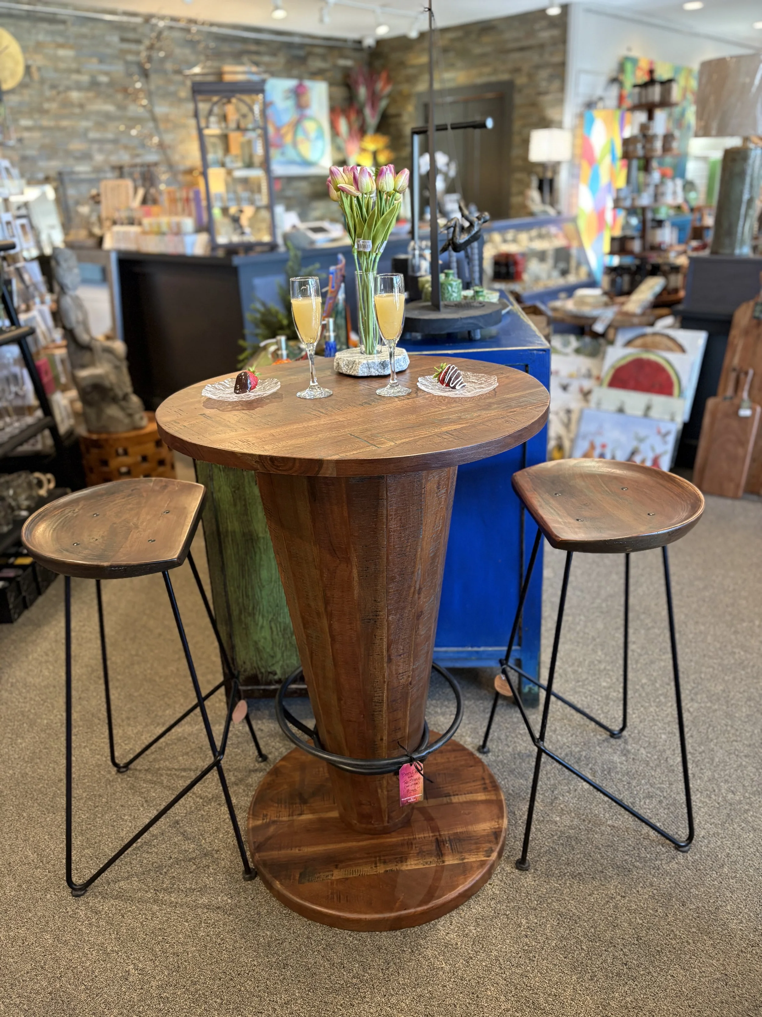A round wooden table with a central pedestal base, decorated with a vase of pink tulips, two champagne glasses filled with a yellow beverage, and two small plates with chocolate-covered strawberries. The setting appears to be in an art or thrift stor