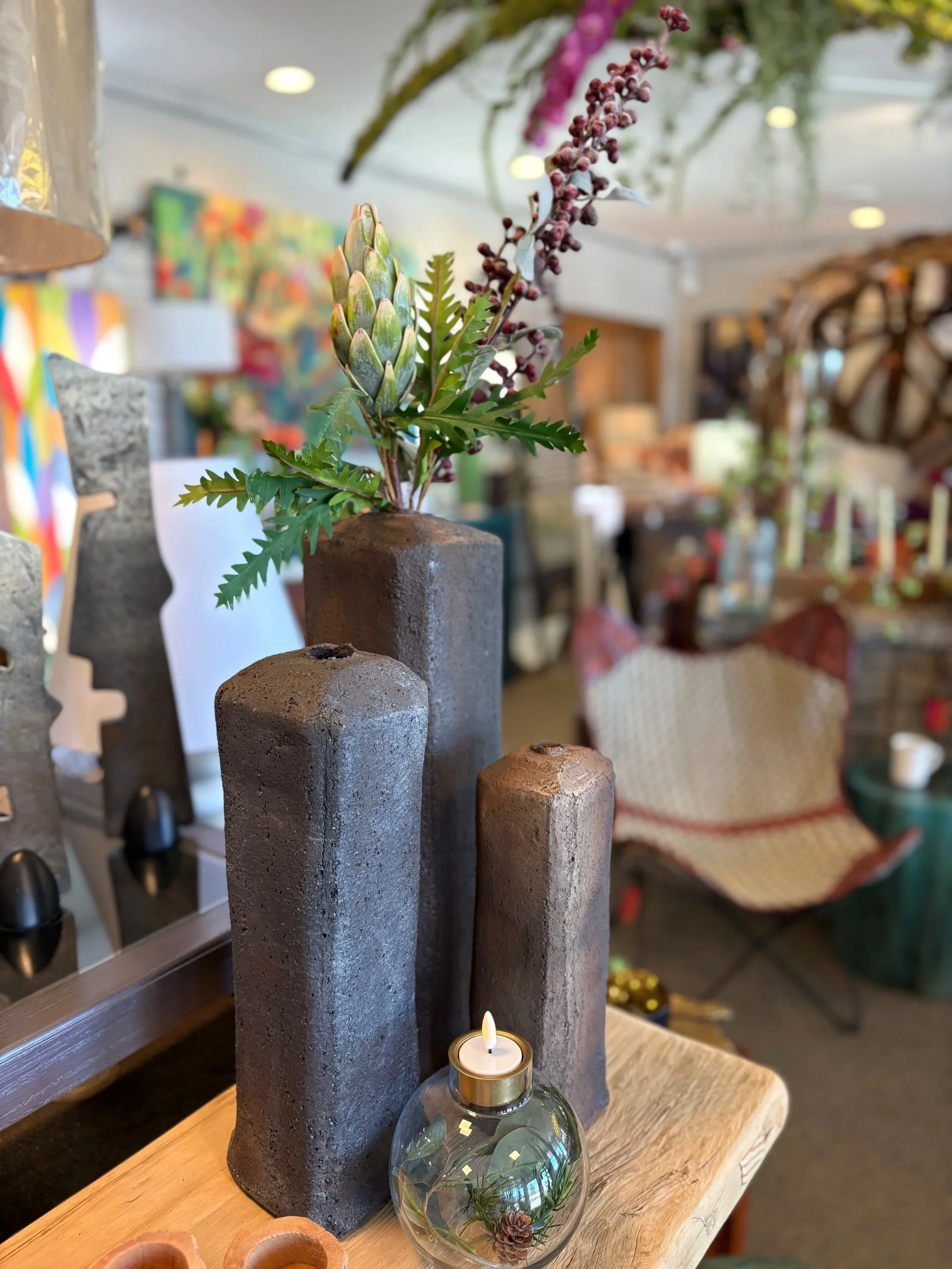 Decorative arrangement of natural elements including an artichoke, red berries, and fern leaves in a textured vase, with a lit candle in a glass holder in front, set on a wooden surface. In the background, colorful artwork and indoor furnishings are visible.