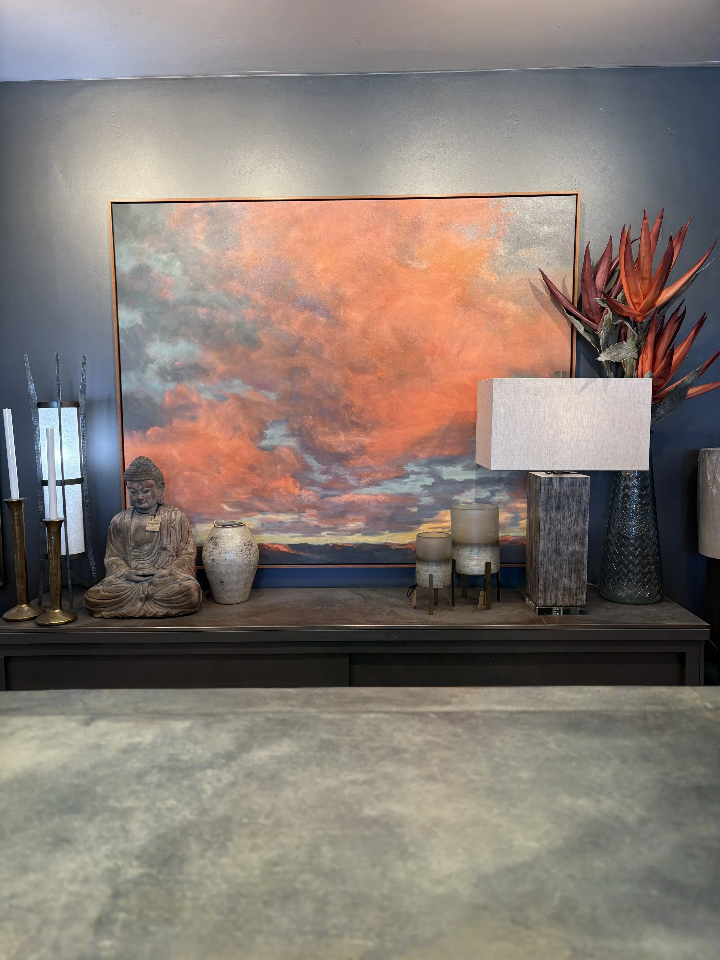 A display of home decor items, including a large sunset-themed painting, a statue of Buddha, vases, candles, a tall lamp, a large floral arrangement in a vase, and a small table with decorative objects, set against a painted dark blue wall.