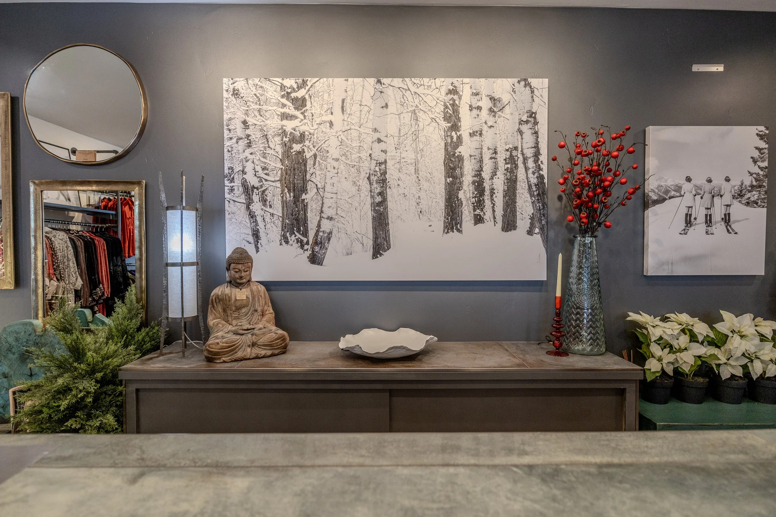 Decorative display with a large black and white forest picture, a red berry branch in a tall vase, a Buddha statue, and various other decorative items on a console table, against a gray wall.