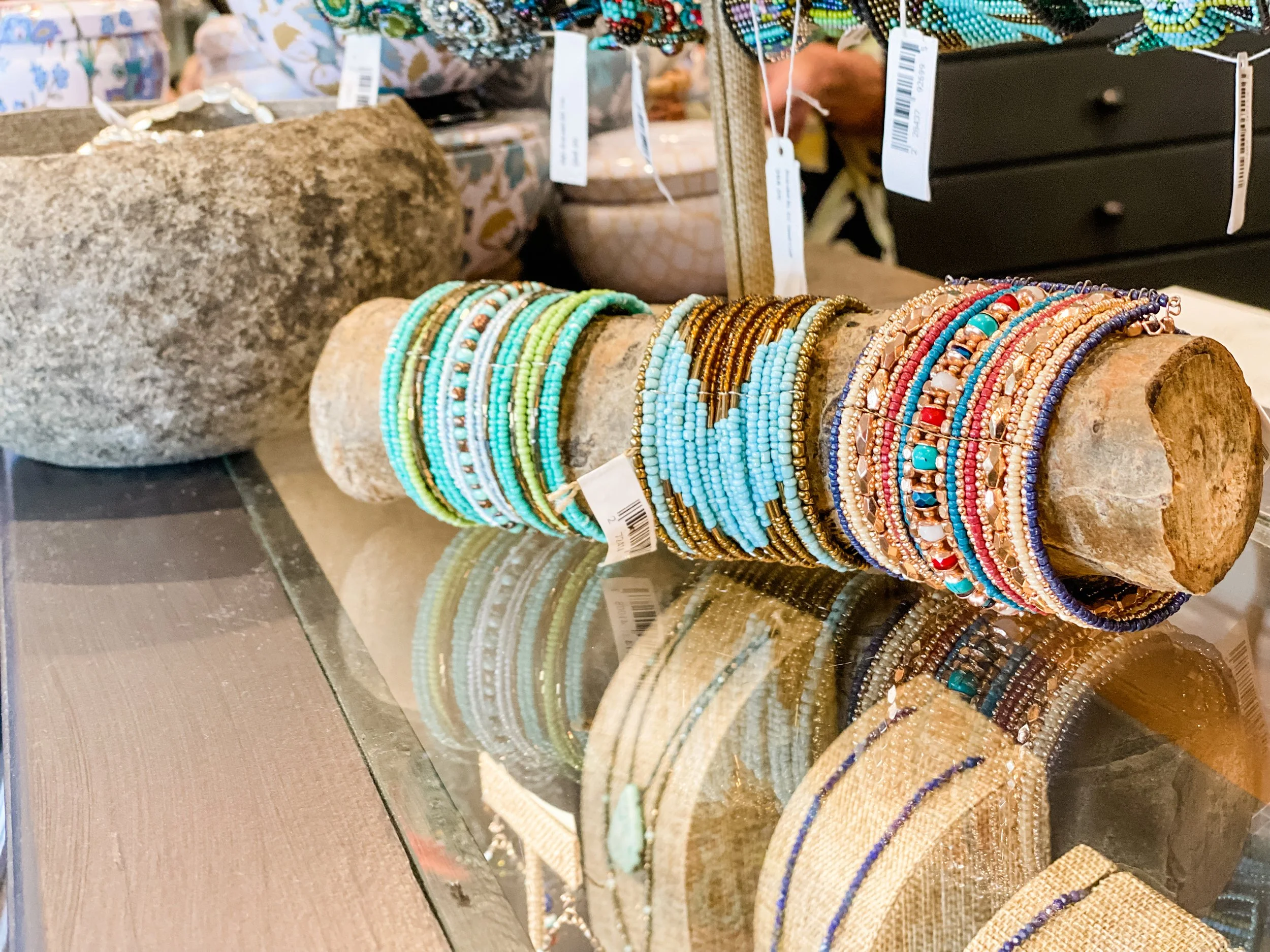 Colorful beaded bracelets displayed on a rustic wooden log at an outdoor market stall, with additional accessories and tags visible in the background.