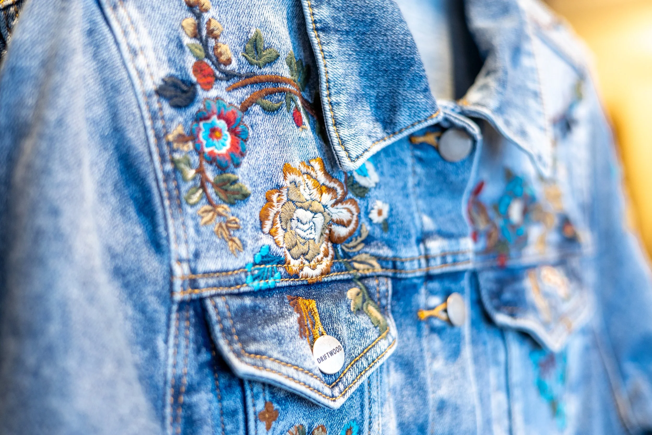 Close-up of a denim jacket with colorful floral embroidery, button closure, and a small white label reading 'DRIFTWOOD'.