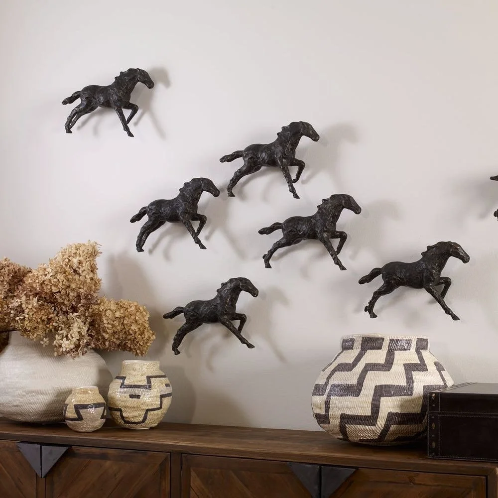 Decorative wall art with six black horse sculptures in a jumping pose arranged in a pattern. The wall is white, and there are ceramic vases and dried flowers on a wooden surface below the art.
