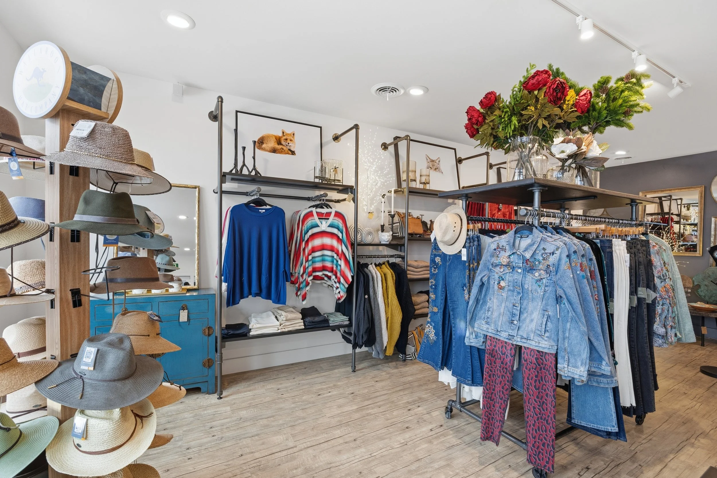Clothing store display with hats on a wooden stand, shirts, jeans, and a large floral arrangement on a table.