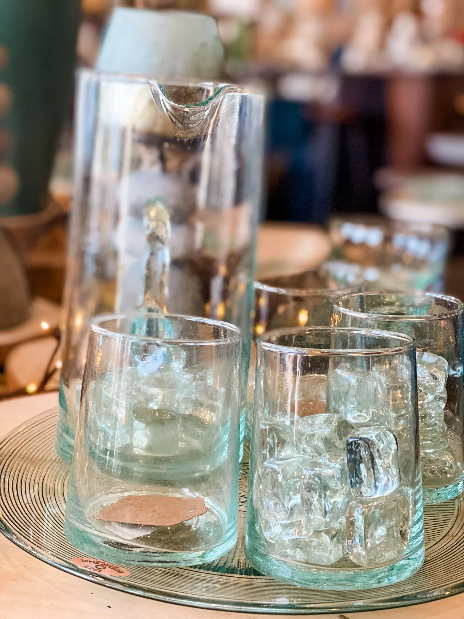 Close-up of multiple clear glass mugs with handles, arranged on a round decorative tray or plate, with a blurred background of a restaurant or cafe setting.