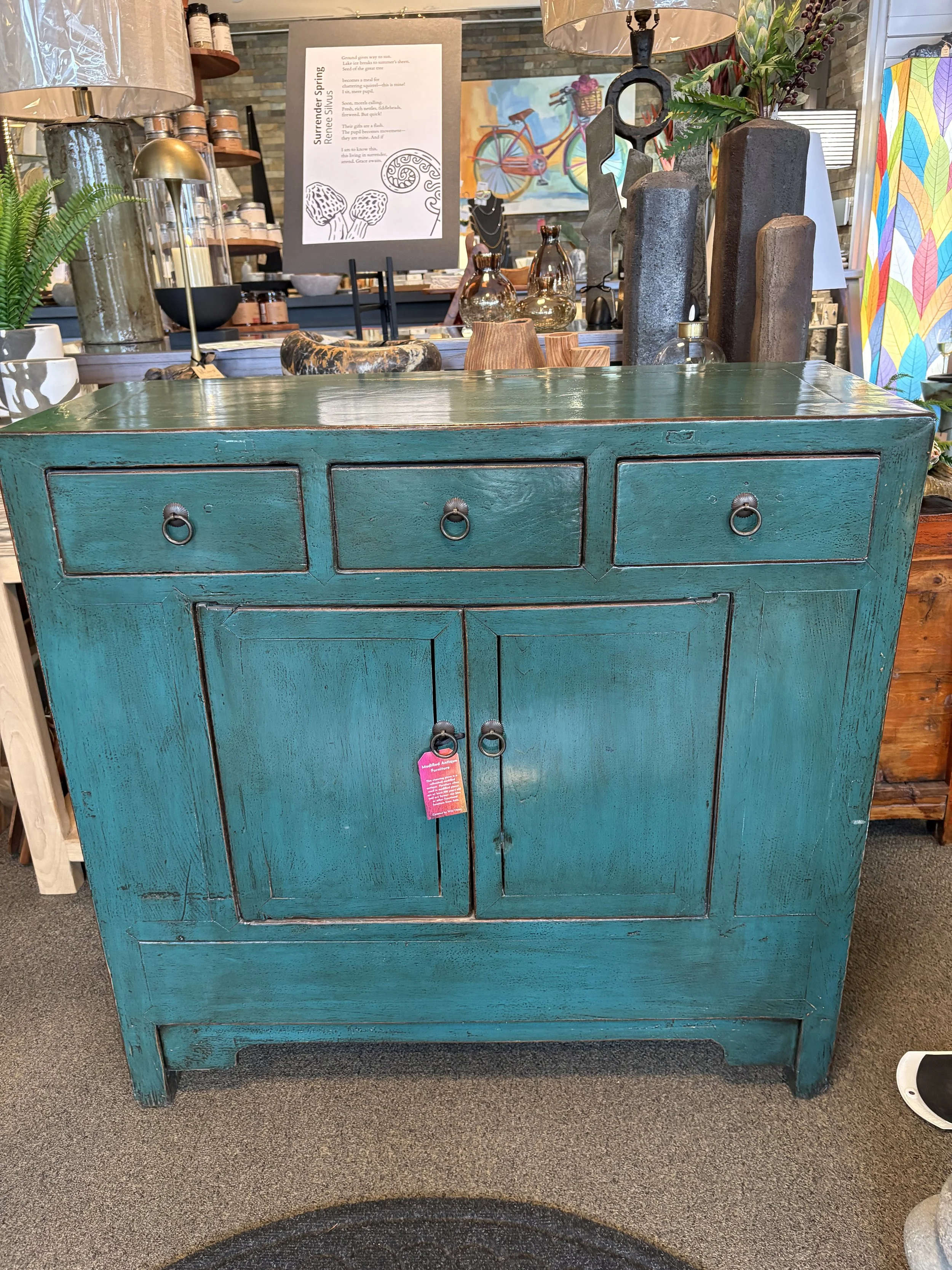 A teal-colored antique wooden cabinet with three small drawers on the top row and two larger doors below, set on a gray carpeted floor in a store. The cabinet has round metal ring pulls on the drawers and doors, with a pink gift tag hanging from the 