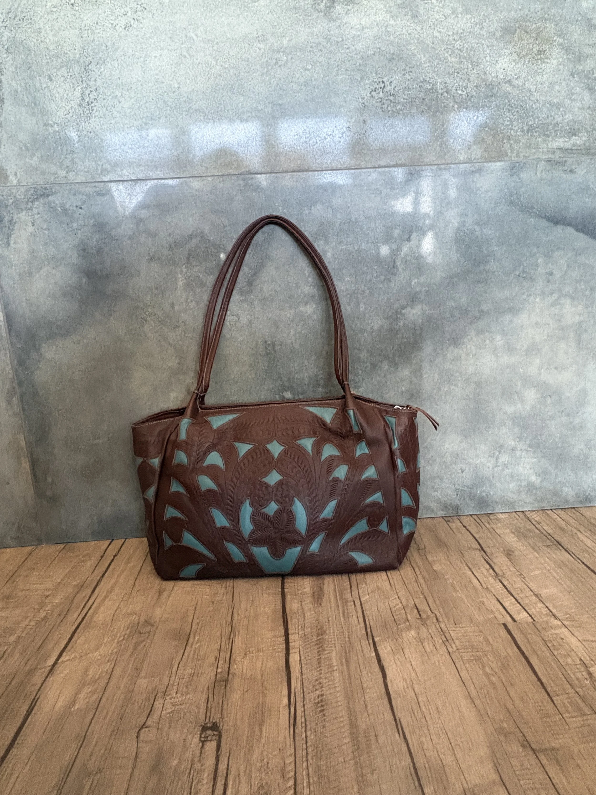 A brown leather handbag with intricate cut-out patterns and teal accents, resting on a wooden floor against a gray textured wall.