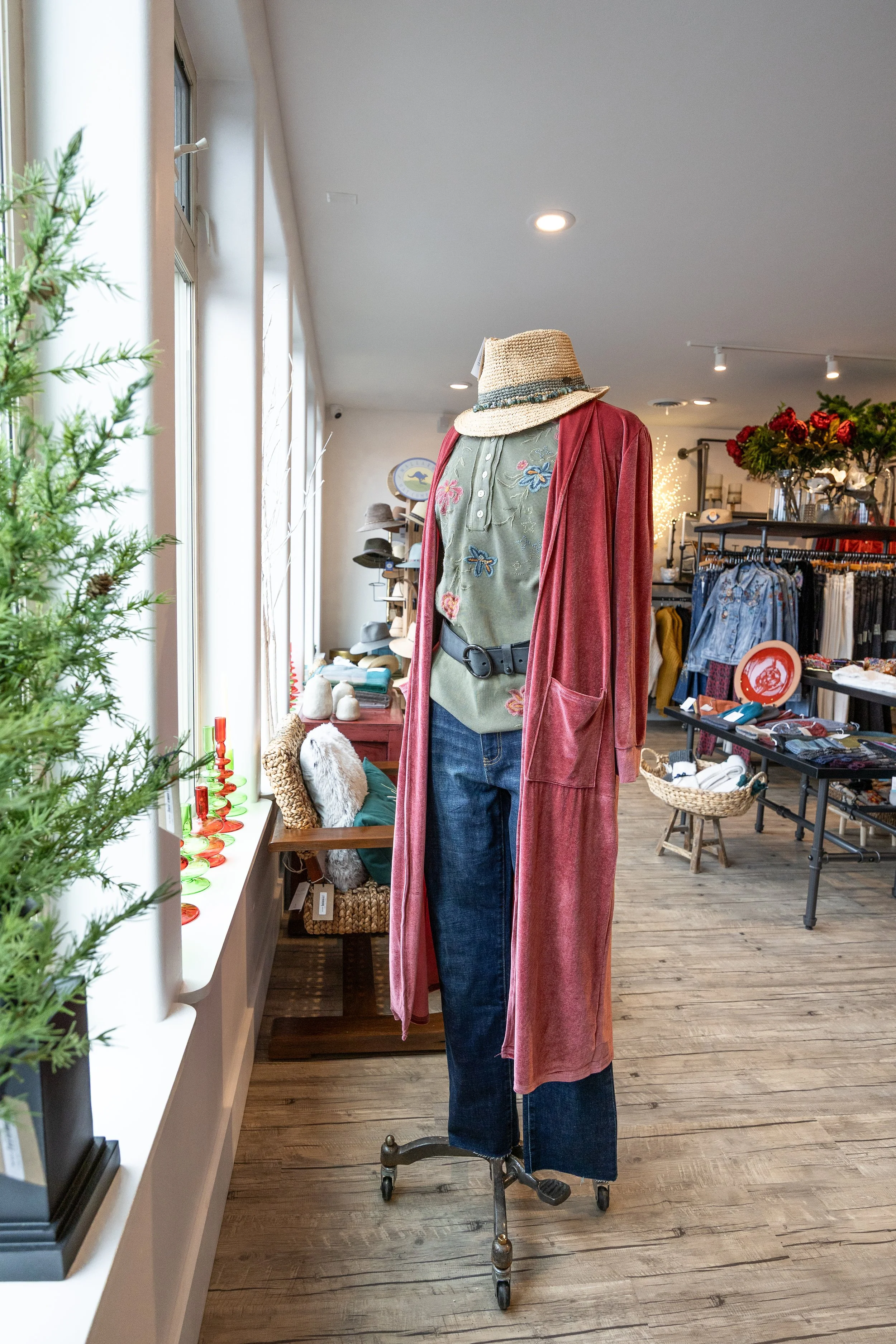 Display of clothing on a mannequin in a boutique, featuring a straw hat, green embroidered top, red cardigan, and dark jeans. Store interior with accessories, hats, and clothing racks visible.