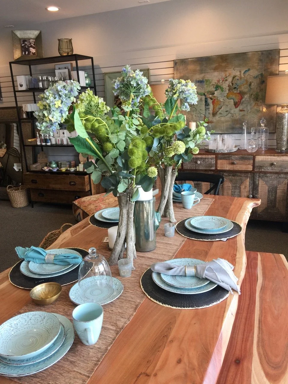 A dining table set for six, with decorative plates, cups, and napkins, accented by a large floral centerpiece of hydrangeas in a vase, in a cozy, inviting living space.