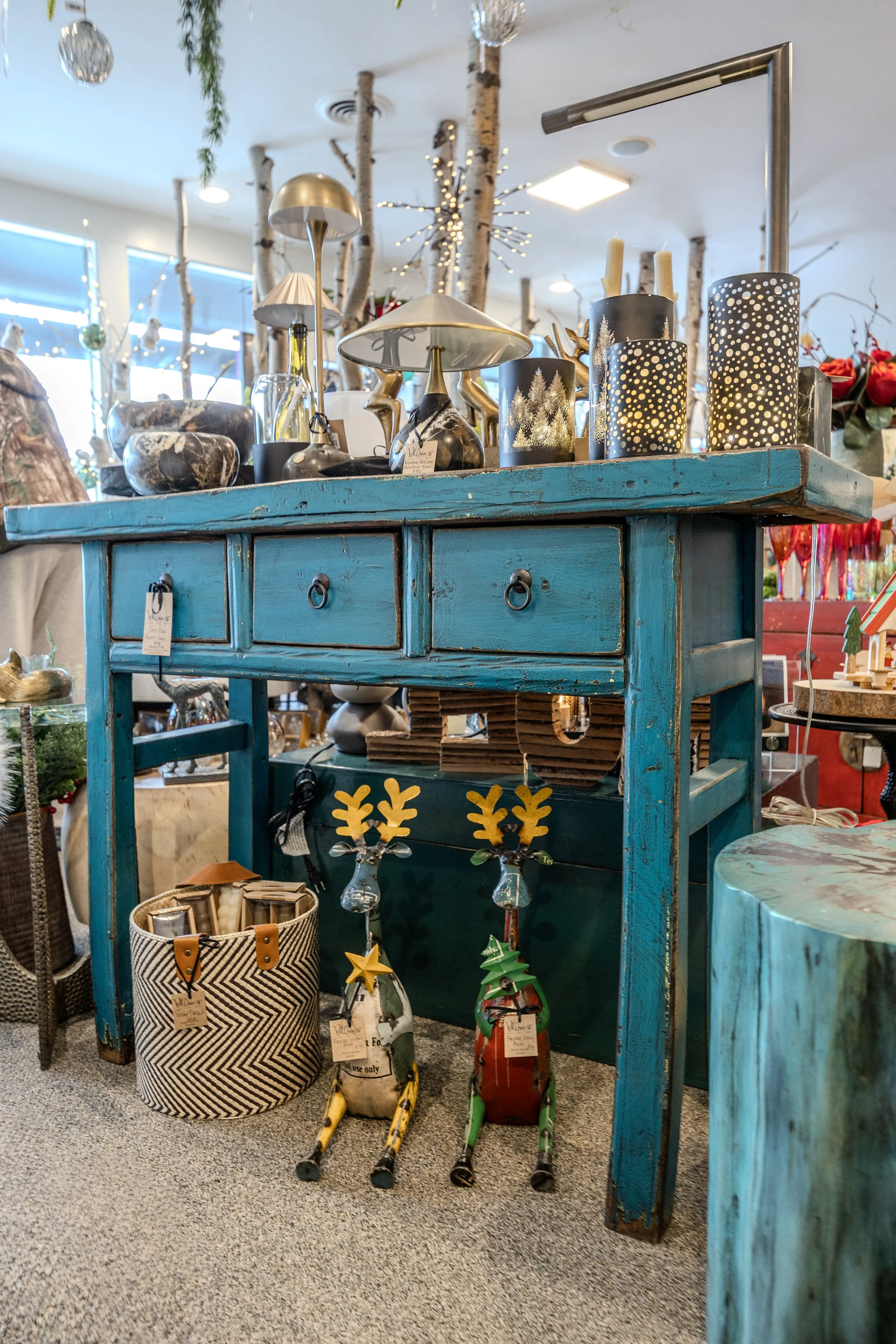 Display of holiday-themed decorative items and accessories including three pairs of colorful miniature skis, candle holders with candles, and various other decorations on a distressed blue wooden table. In the background, there are decorative tree branches with lights and ornaments, and sunlight coming through window blinds.