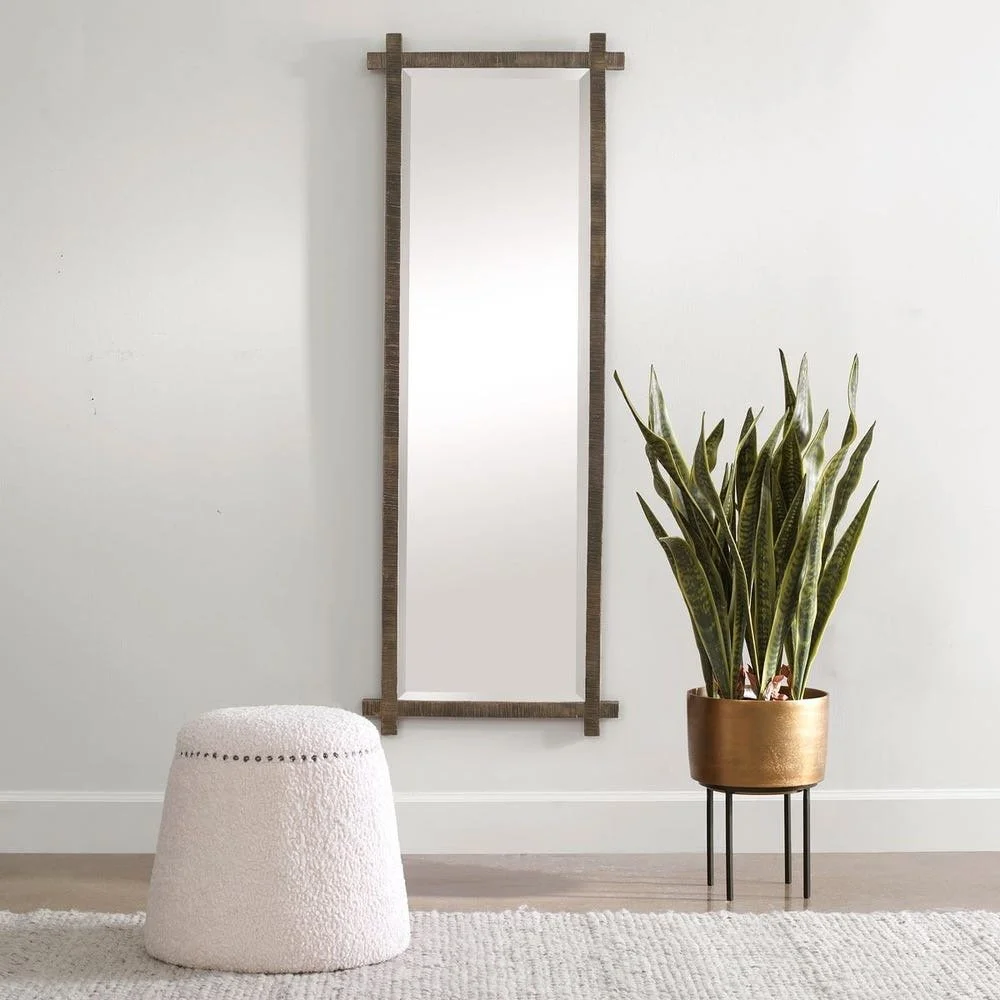 A tall, rectangular wall mirror with a wooden frame hanging on a plain white wall. To the right of the mirror, there is a large potted snake plant in a gold-colored planter on a black plant stand. To the left of the plant, there is a small, round, white textured stool and a light-colored area rug on the floor.