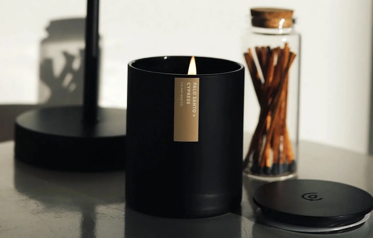 A lit black candle labeled 'Palo Santo + Cypress' on a table, with a black circular object, a mirror, and a glass container filled with cinnamon sticks in the background.