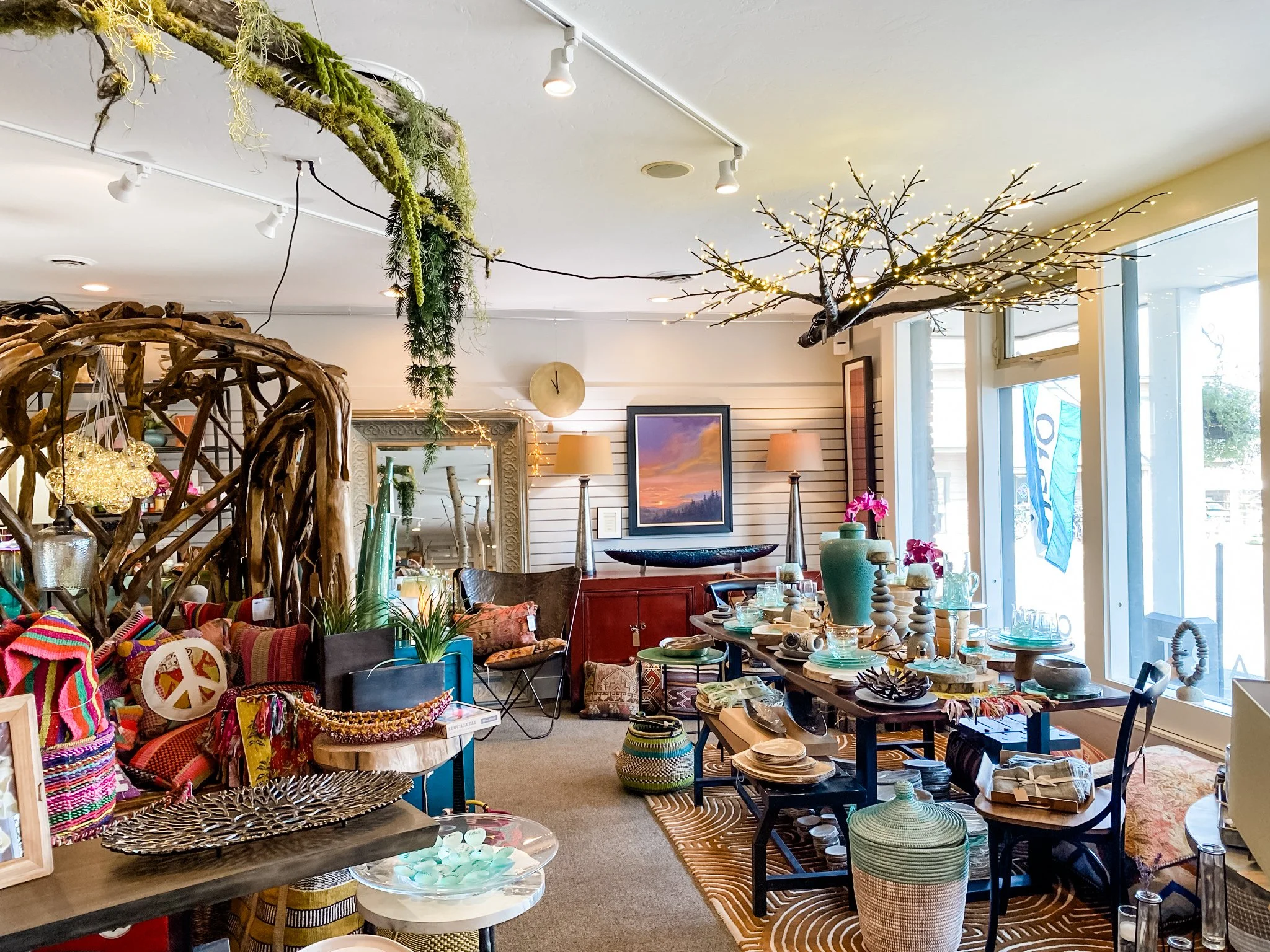 Interior of a store or gallery with various decorative items, including colorful woven textiles, vases, pottery, furniture, and decorative branches with lights, arranged in a vibrant and eclectic style.