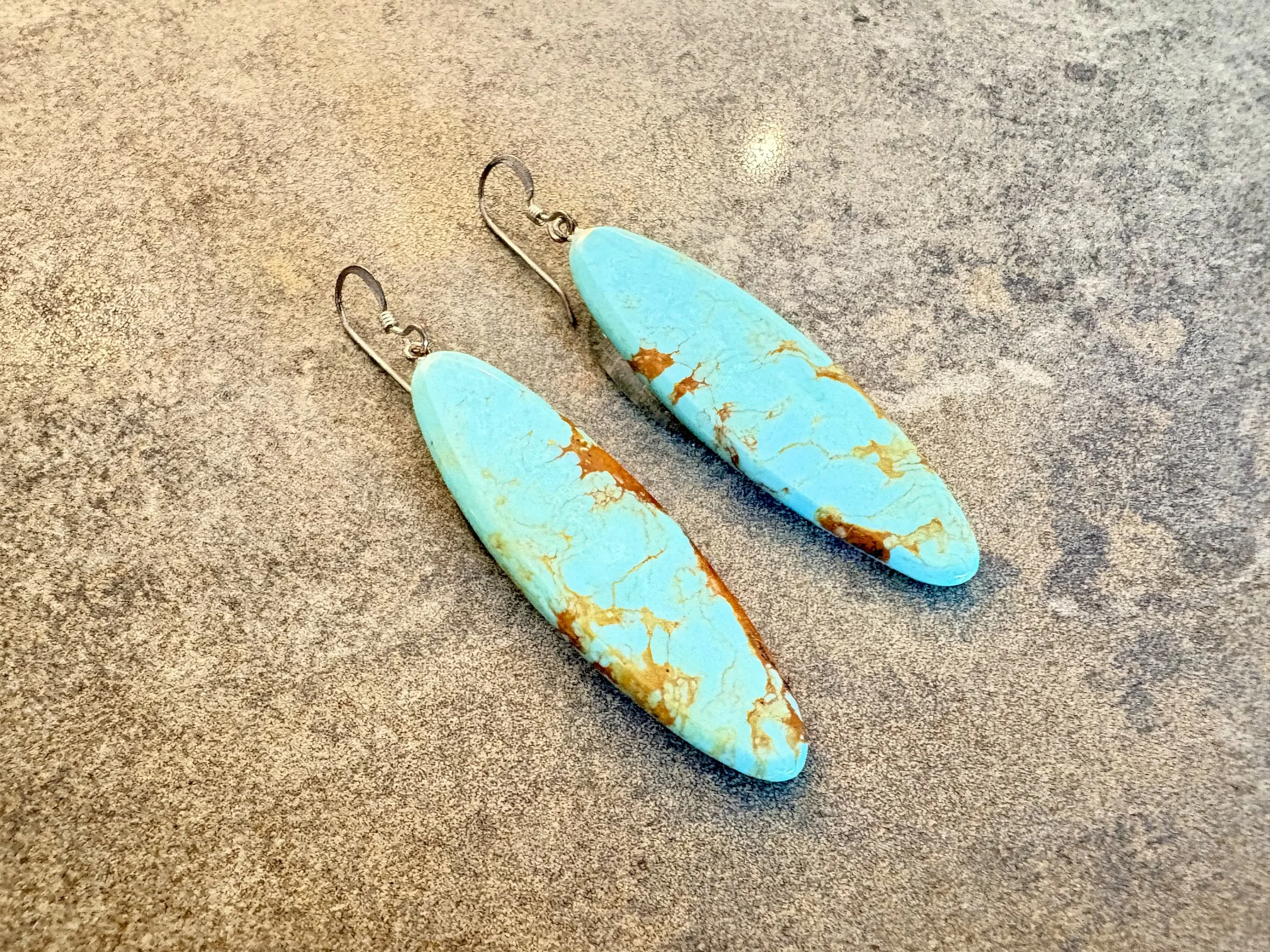 A pair of turquoise and orange stone earrings lying on a speckled gray surface.