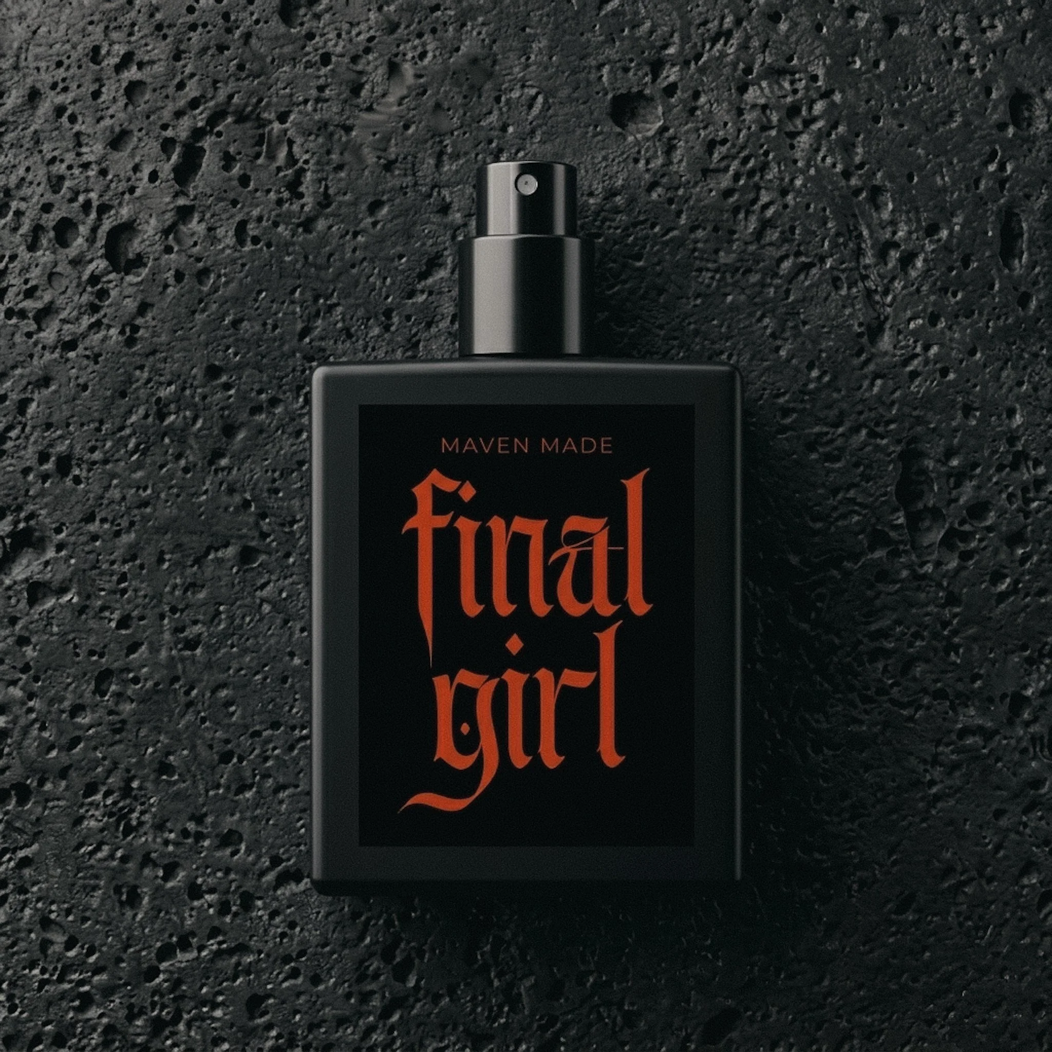Flat lay overhead view of Maven Made 'Final Girl' perfume bottle on textured black rock. The matte black bottle features bold red gothic text.