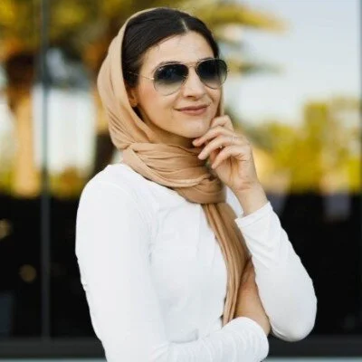 A woman wearing sunglasses and a beige hijab, smiling and standing outdoors with trees in the background.
