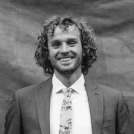 A man with curly hair and a beard wearing a suit and tie, smiling at the camera.
