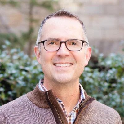 Portrait of a smiling man wearing glasses, a checkered shirt, and a brown sweater, standing outdoors in front of a stone wall and greenery.