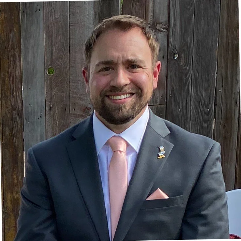 Man smiling in a blue suit with pink tie and pocket square, standing in front of wooden fence.