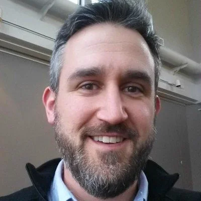 A man with dark hair and a beard smiling, wearing a blue shirt and a black jacket, indoors with a beige wall and a white pipe in the background.