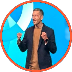 A man standing on stage with a blue background, smiling and gesturing with clenched fists.