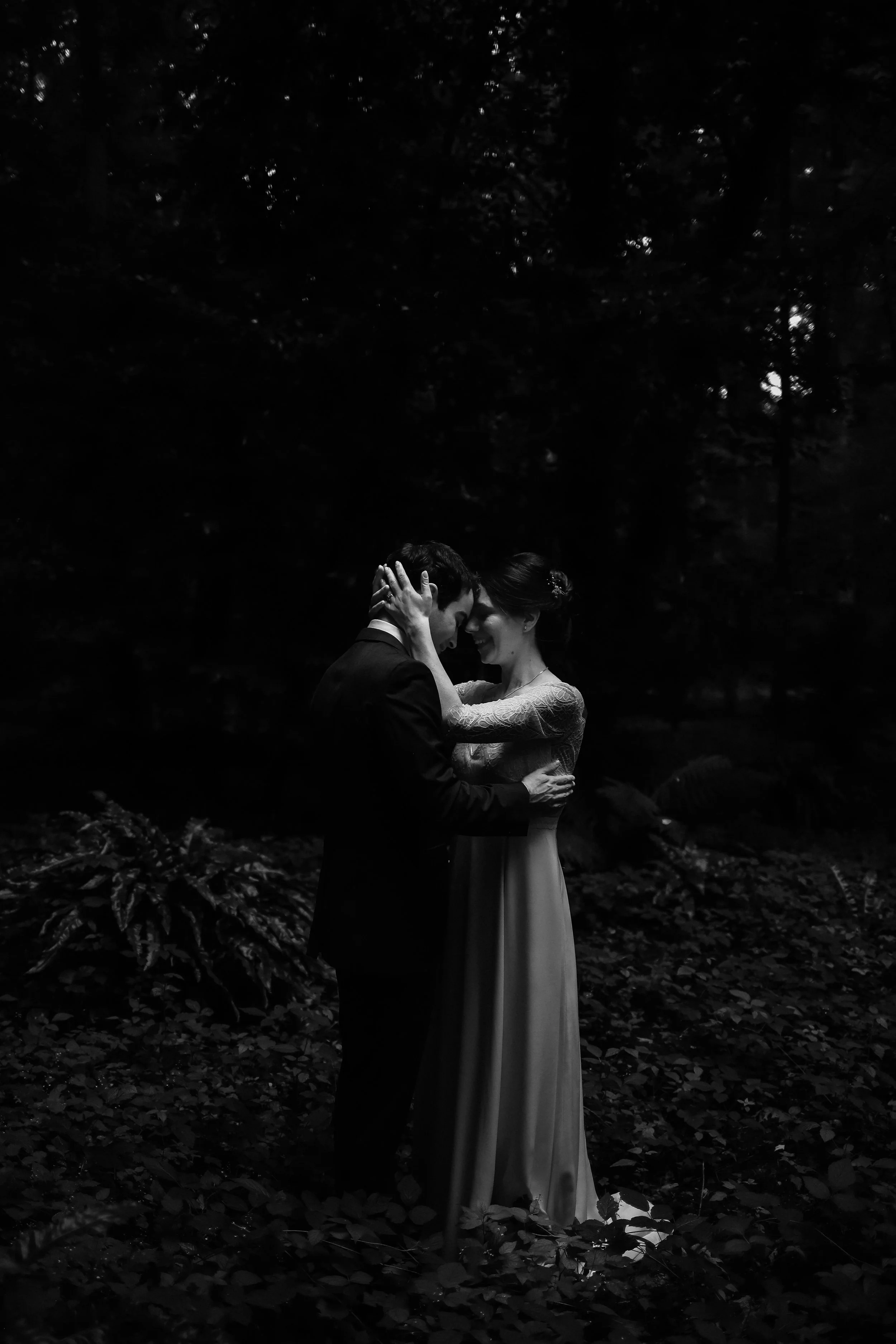 Un couple en robe de mariage s'embrassant dans une forêt sombre, éclairé partiellement par la lumière naturelle.