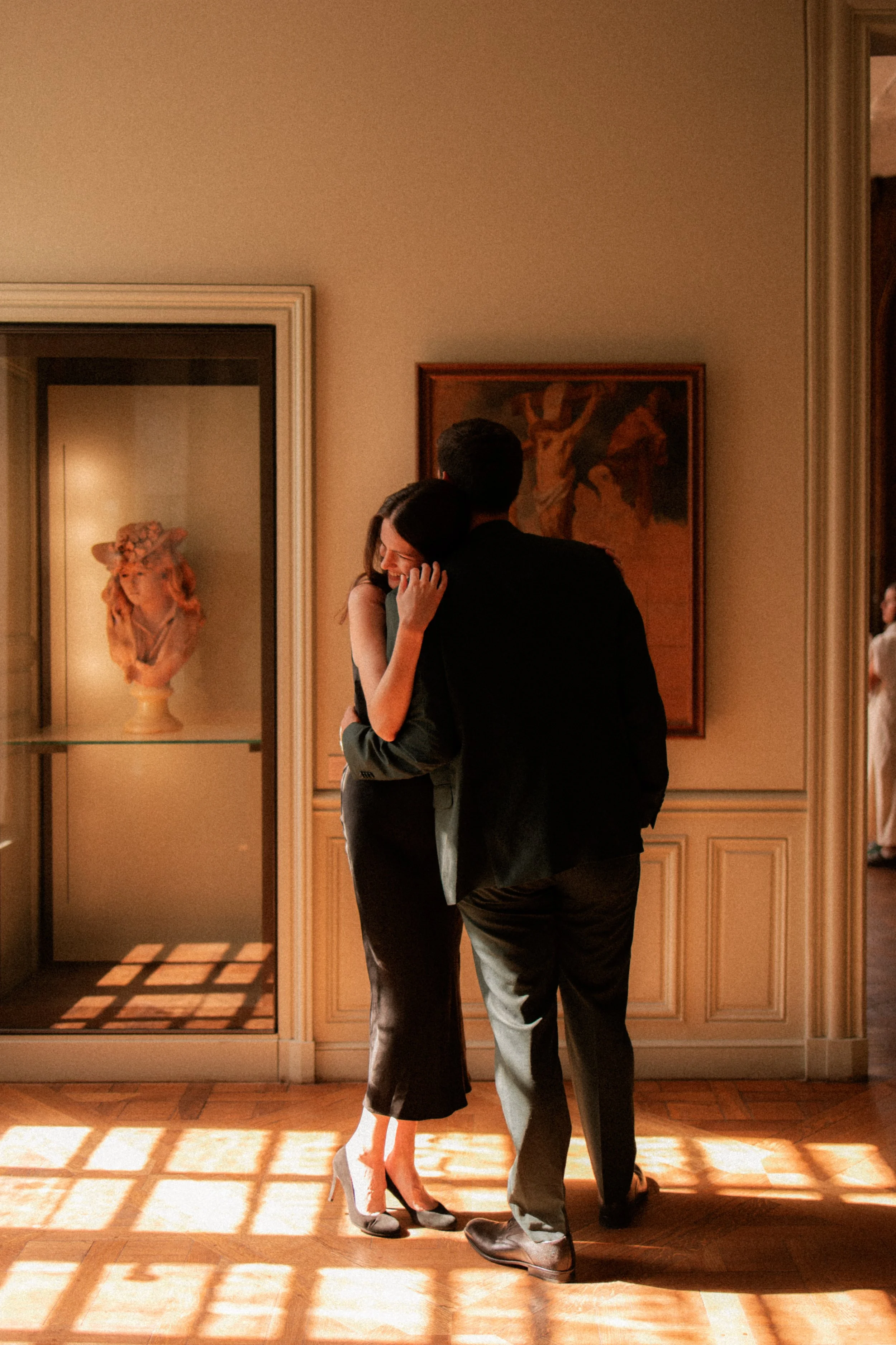 Deux personnes, un homme et une femme, s'embrassent dans une pièce décorée avec des peintures classiques, près d'une vitrine avec une sculpture en céramique. La lumière naturelle illumine la scène à travers une fenêtre.