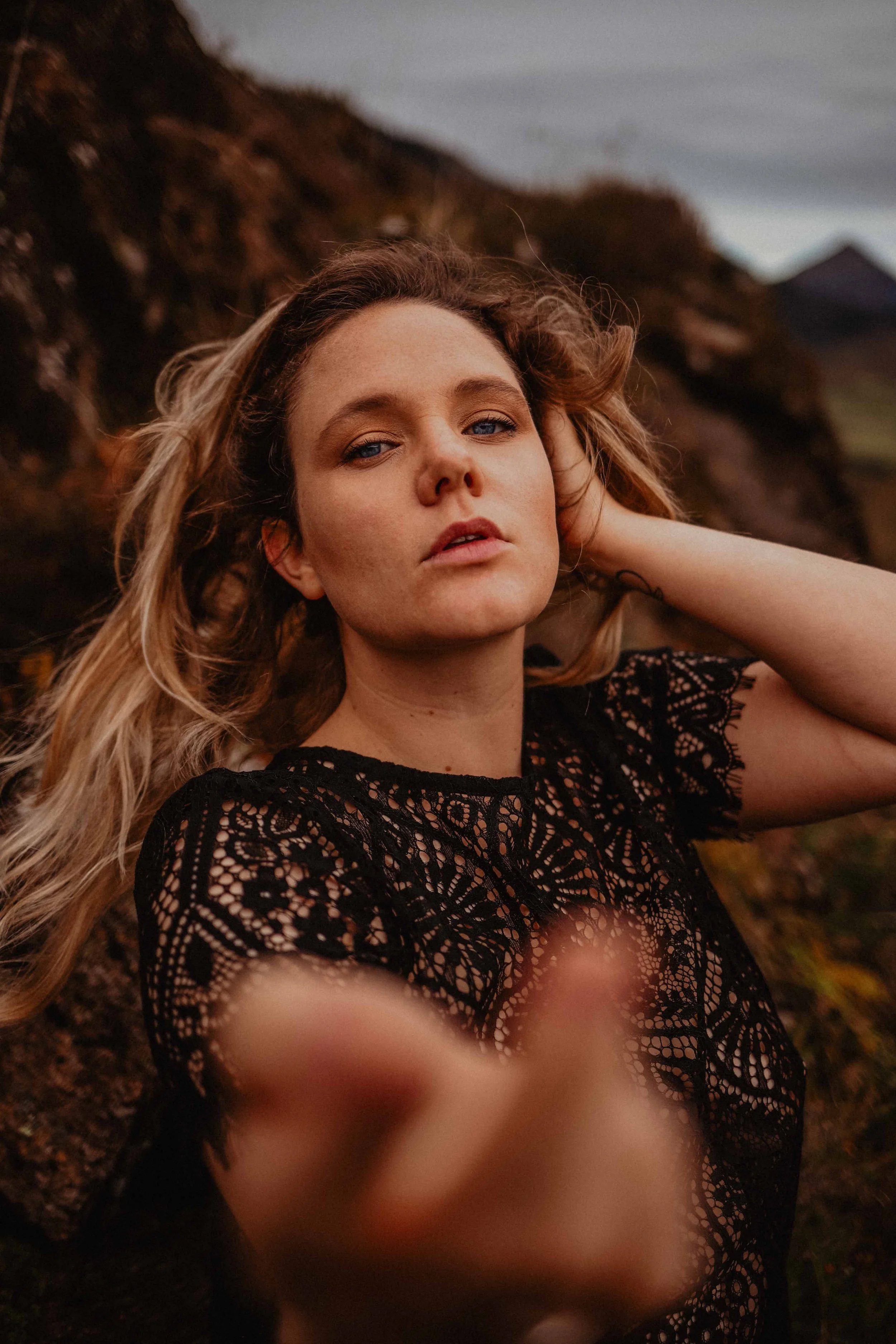 Jeune femme avec des cheveux bouclés portant une robe en dentelle noire, posant de profil avec un fond de paysage naturel rocheux, lynx de des yeux bleus clair, pointant vers la caméra.
