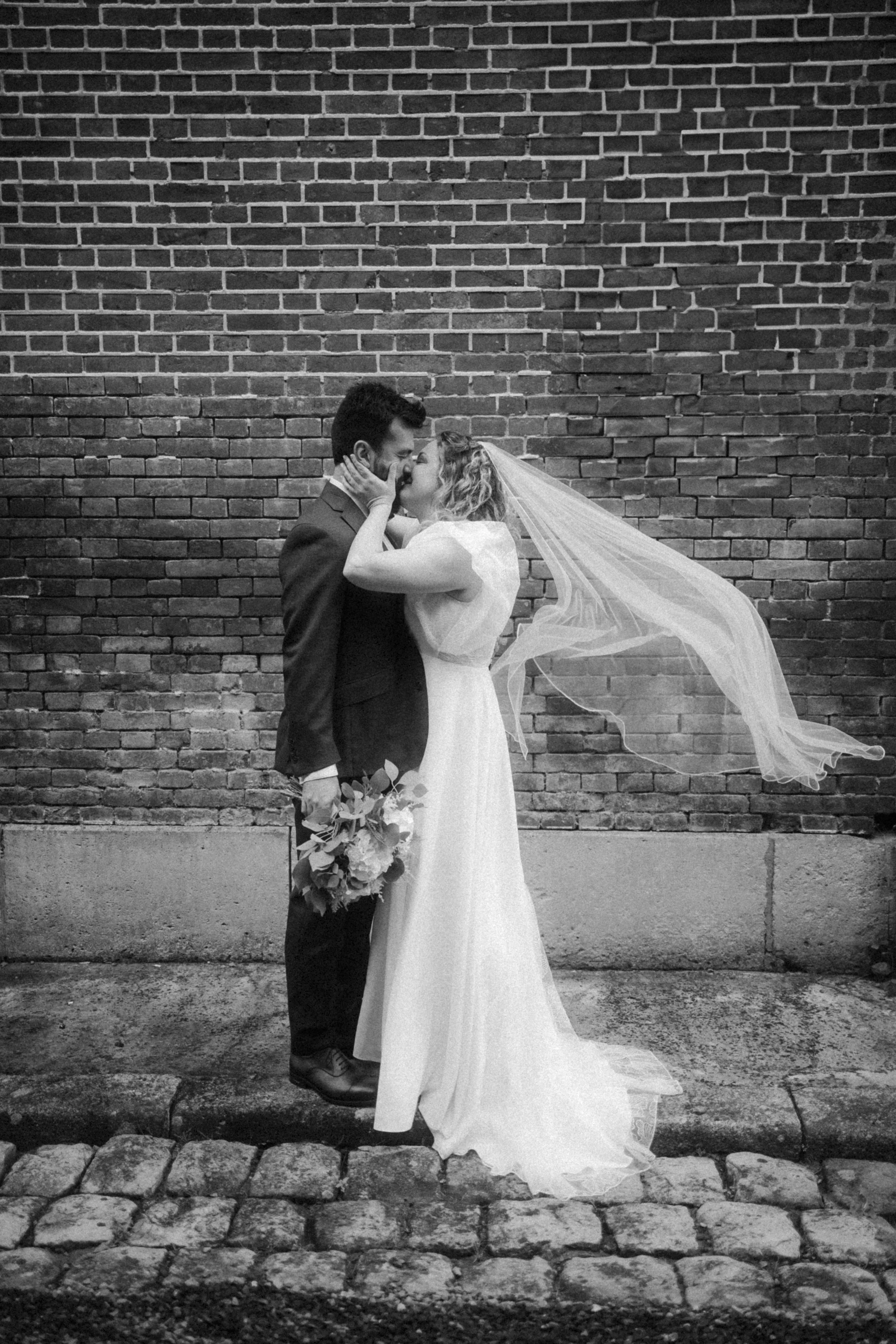 Un couple de mariés s'embrassant devant un mur en briques, la femme portant une robe de mariage et un voile, tenant un bouquet.