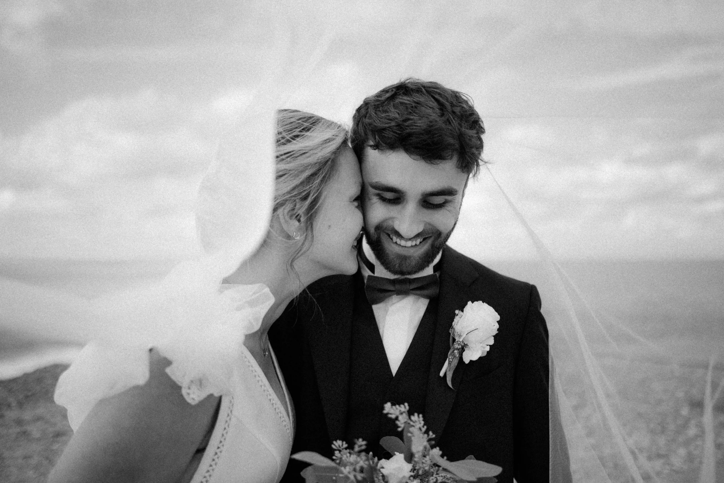 Un couple de mariage souriant, la femme porte une robe blanche et le voile, et l'homme porte un costume noir avec un nœud papillon, dans un paysage naturel.