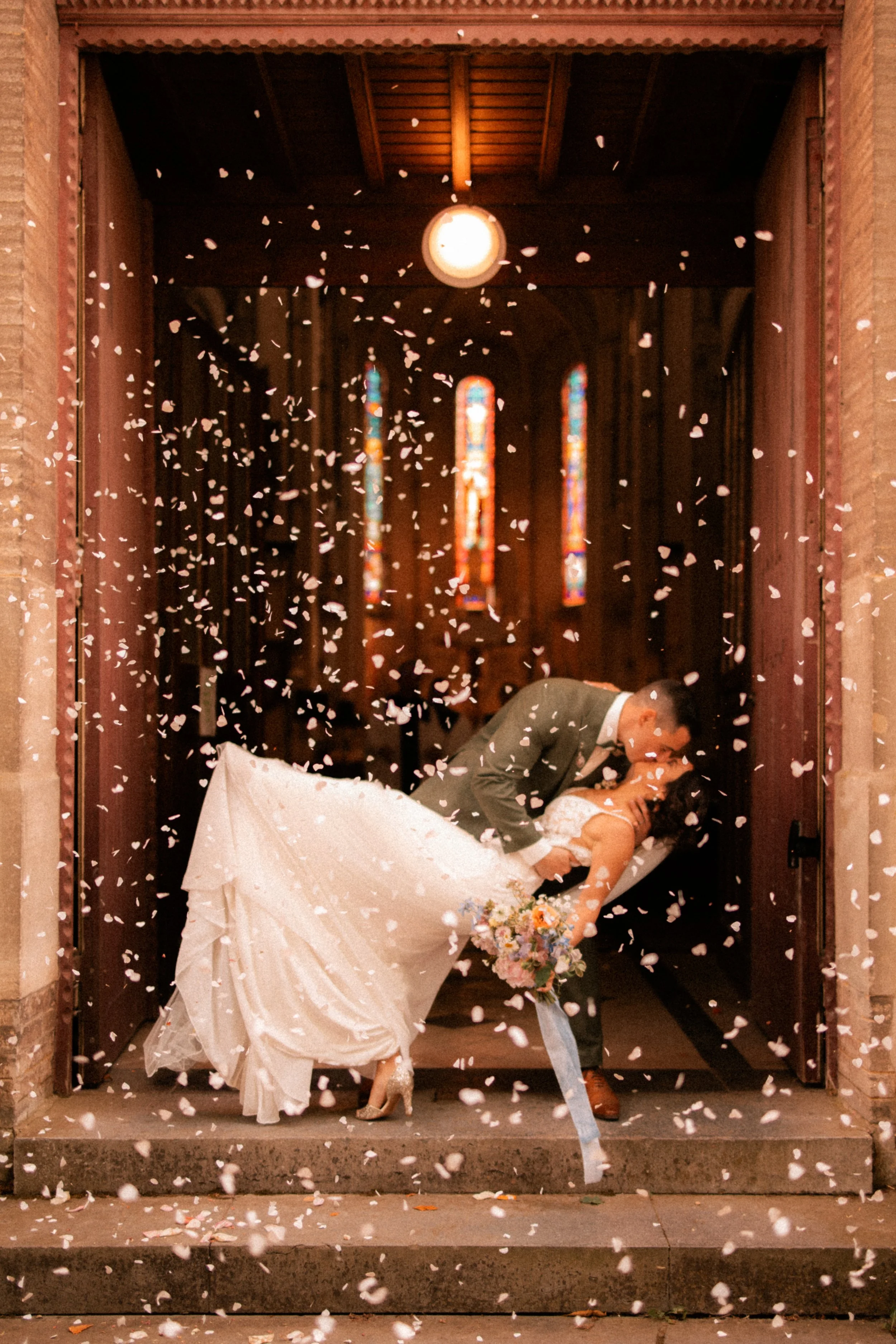 Un couple de mariés s'embrasse devant l'entrée d'une église, avec des pétales feuilles tombant autour d'eux.