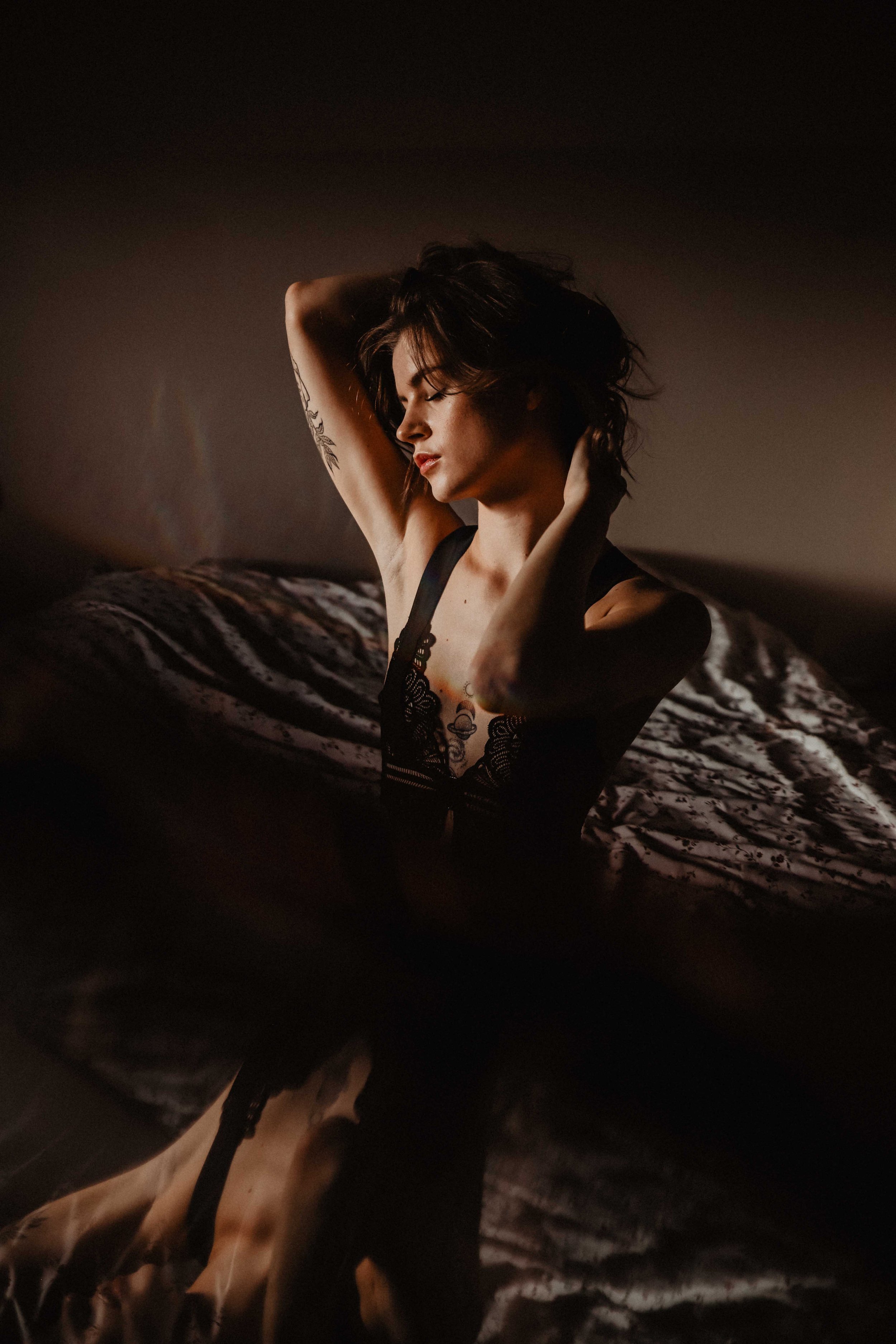 Une femme en lingerie noire pose sur un lit en soulevant ses bras et en touchant ses cheveux, dans une lumière tamisée.