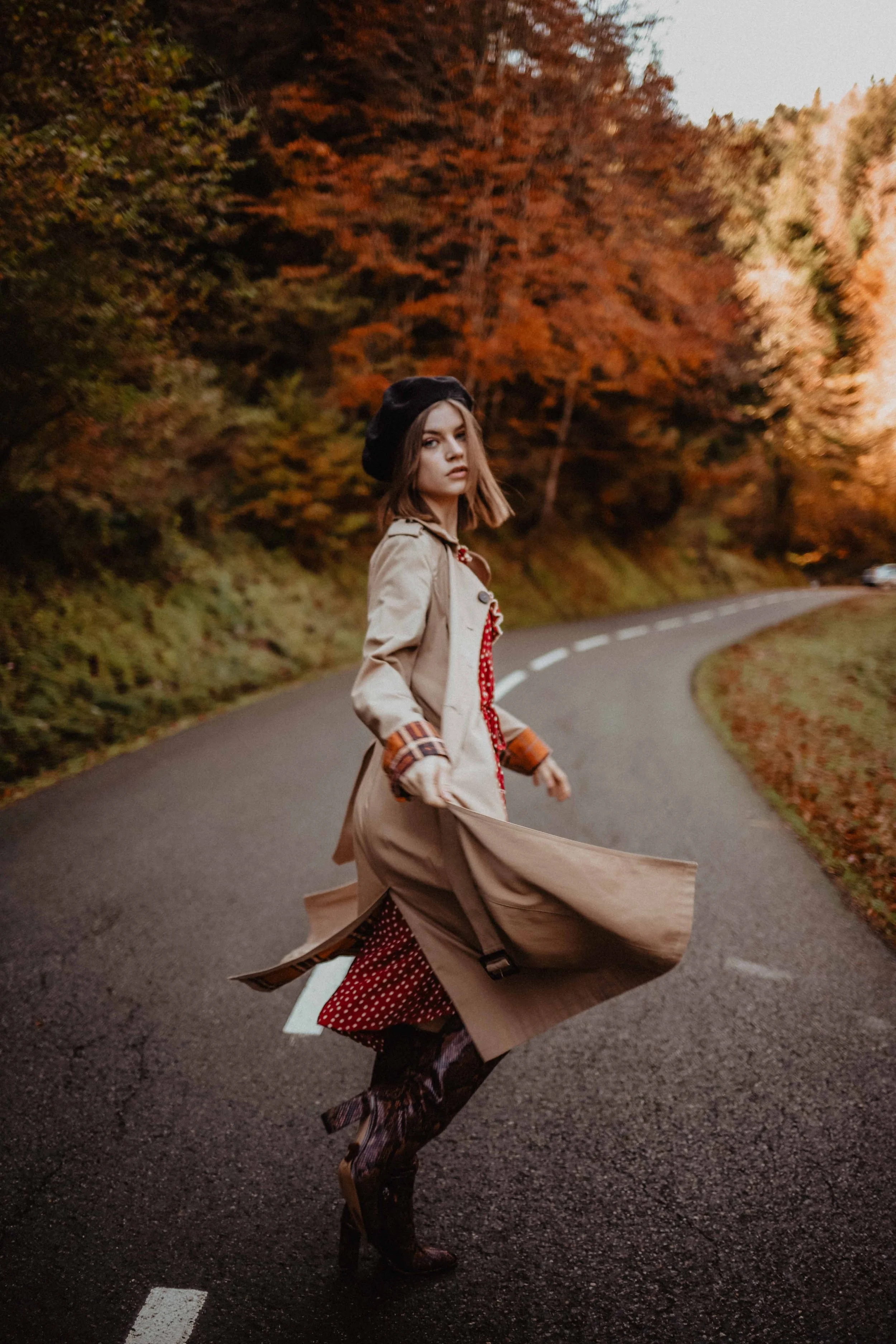 Femme marchant lors d'une journée d'automne, portant un trench beige, un chapeau noir, des chaussures à talons et une jupe rouge à pois, avec un paysage d'arbres aux couleurs d'automne en arrière-plan.