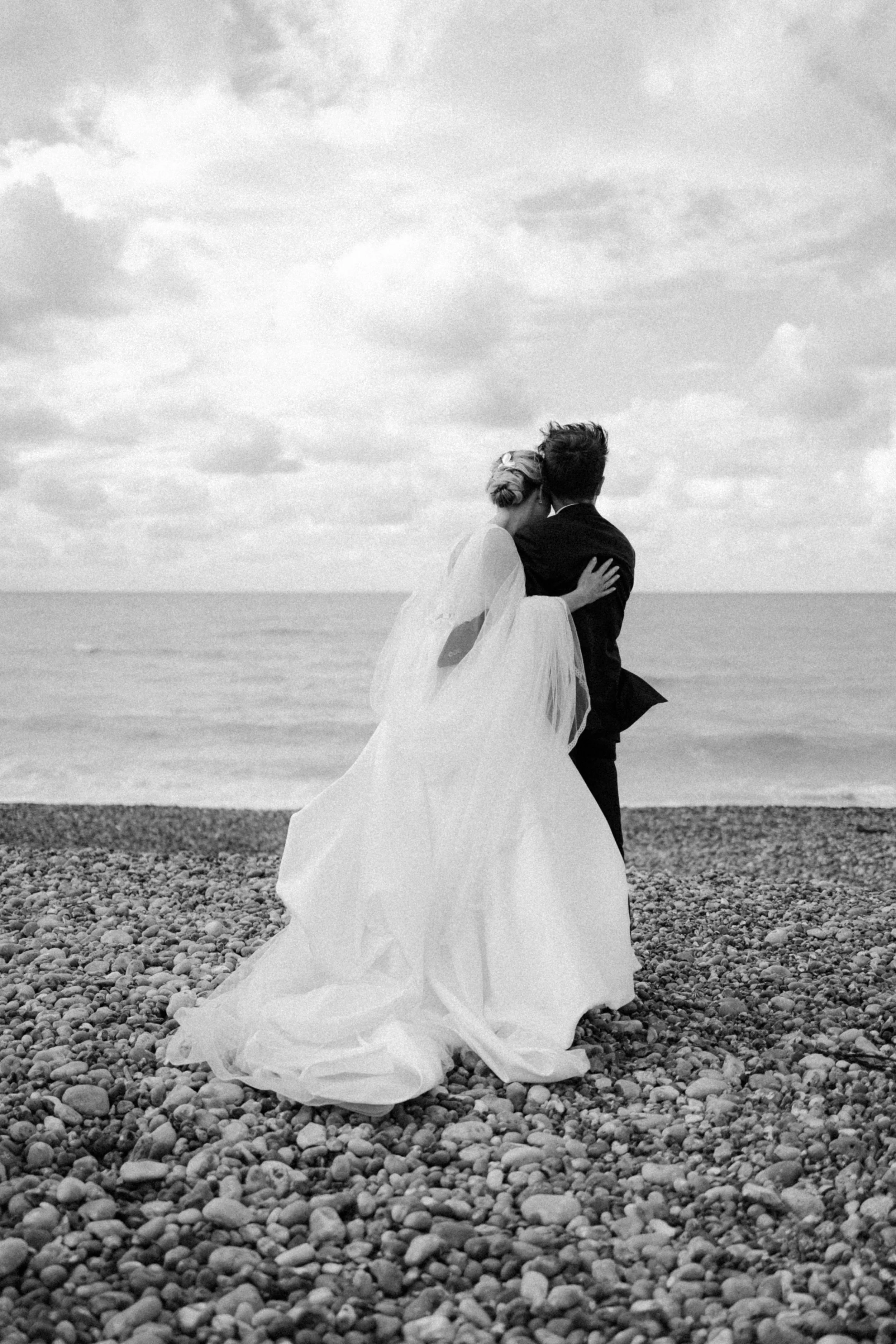Deux personnes en robe de mariage et costume embrassent sur une plage par une journée nuageuse.