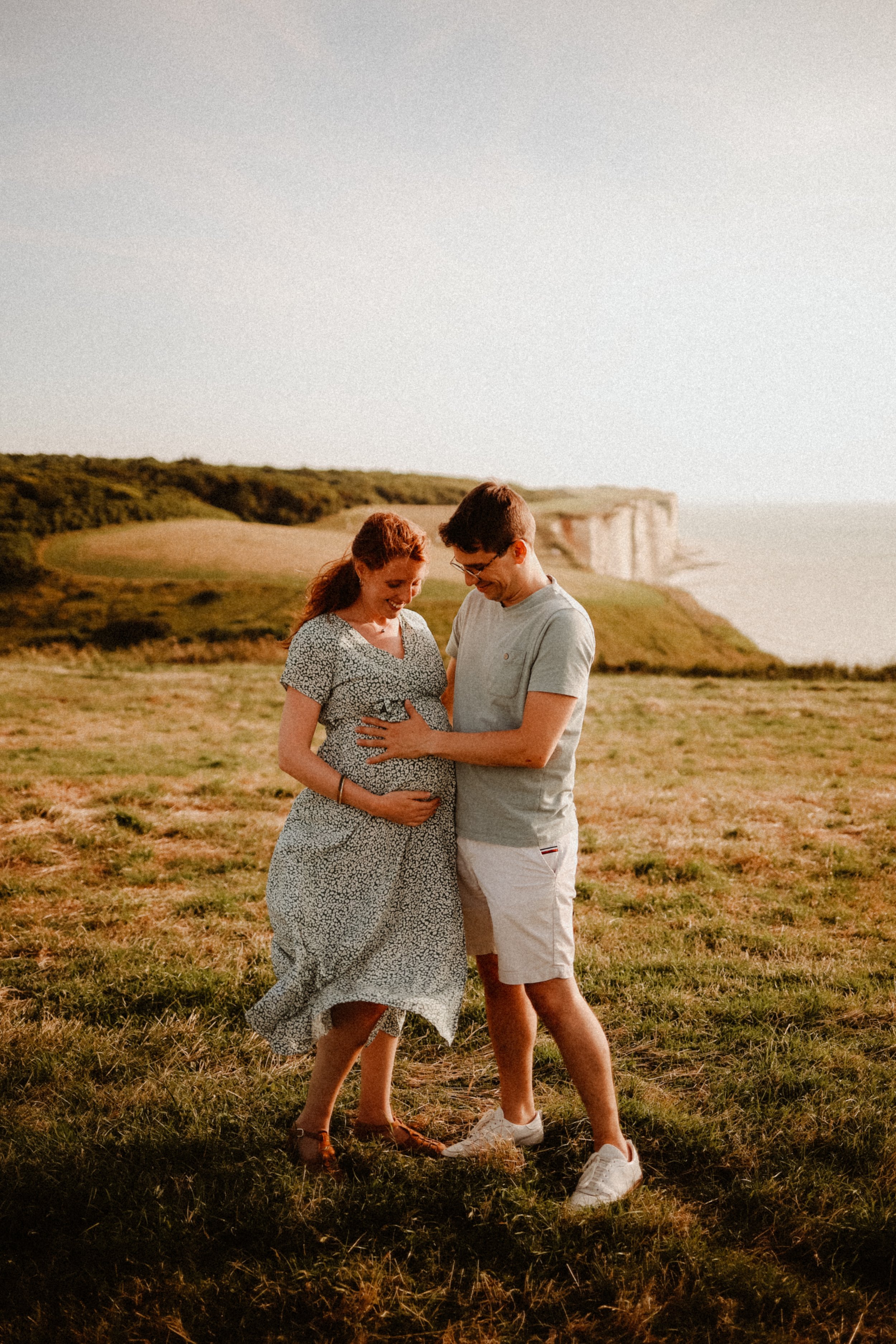 Un couple souriant, la femme enceinte, se tenant la main sur le ventre dans un champ à l'extérieur au coucher du soleil, avec des falaises en arrière-plan.