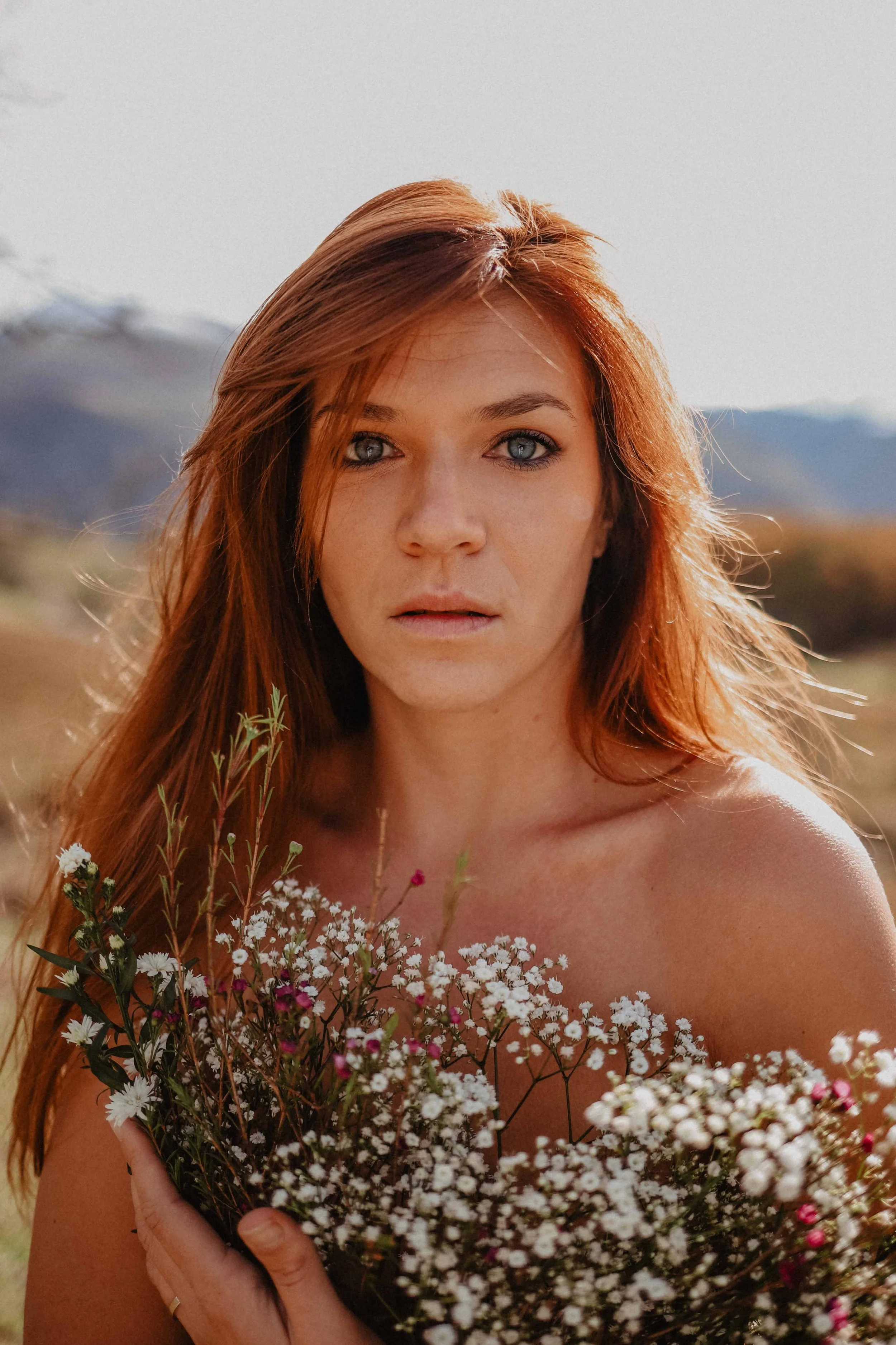 Une femme aux cheveux roux tient un bouquet de fleurs blanches et roses dans un champ, avec des montagnes en arrière-plan.