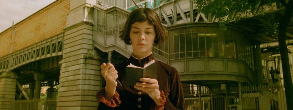 Une femme lit un carnet devant un bâtiment ancien en fer forgé, avec des arbres en arrière-plan.
