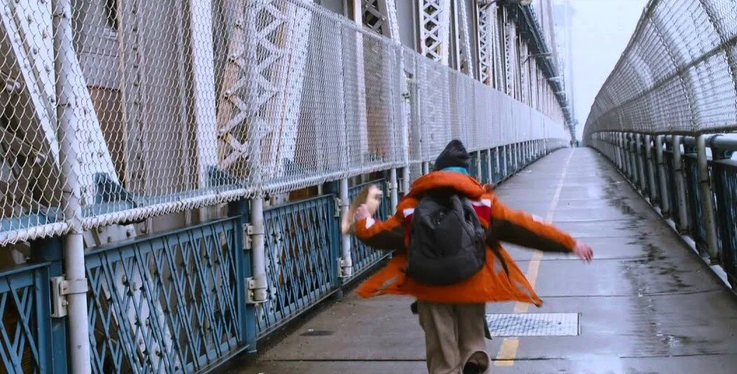 Une personne avec un sac à dos marche sur un pont suspendu, par temps pluvieux, portant un manteau orange et une cagoule noire.