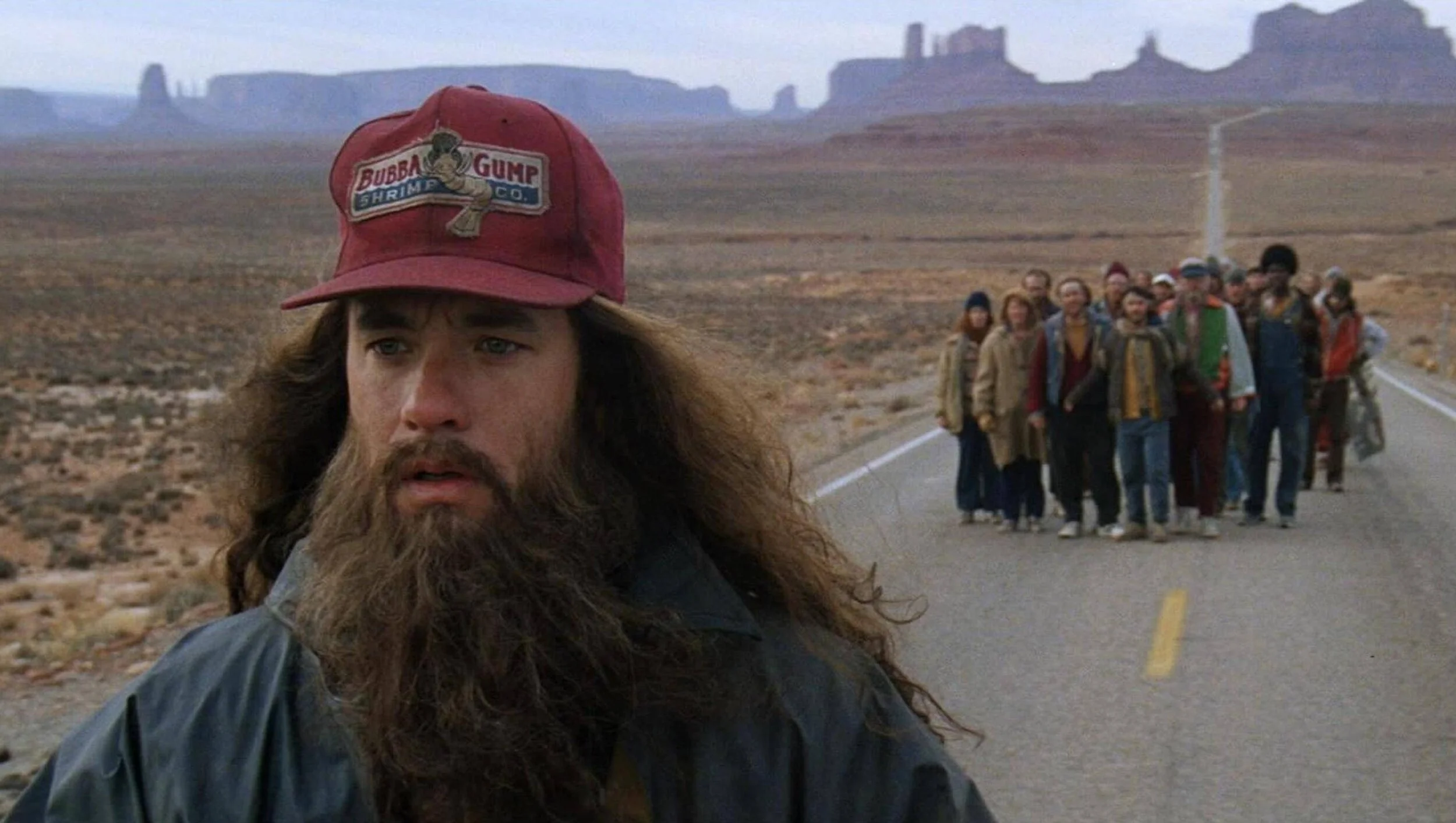 Une personne avec une barbe, portant une casquette rouge avec un personnage de la bande dessinée Bubba Gump et un groupe de personnes marchant dans un désert.