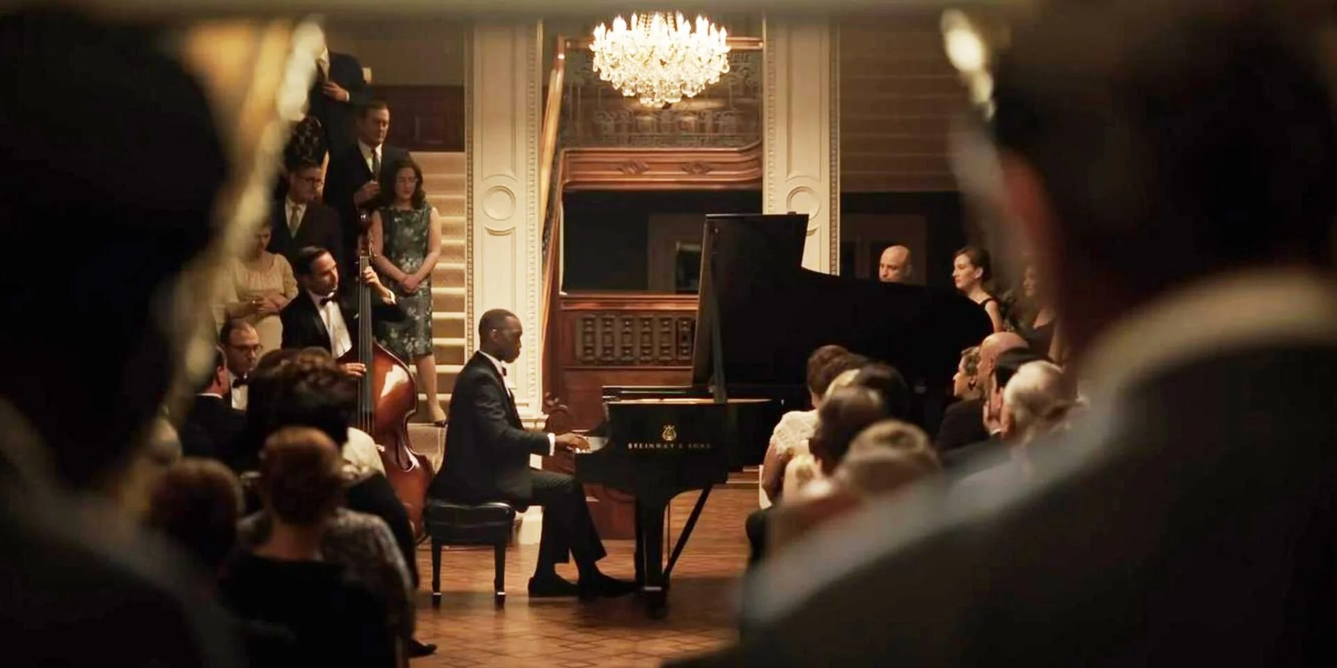 Un pianiste joue du piano à queue dans une salle de concert élégante, entouré de musiciens et de spectateurs, avec un lustre chandelier au plafond.