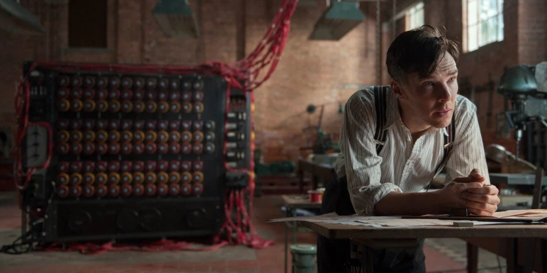 Un homme jeune avec une coiffure rétro, portant une chemise à manches longues rayée, penché sur une table en bois, dans un atelier lumineux en briques avec des outils et matériaux.