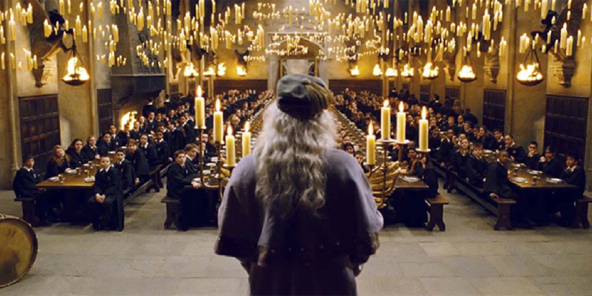 Un groupe de personnes en robes noires assises à une longue table dans une salle ornée de chandeliers et de bougies, avec un personnage en costume de rat placé au centre en premier plan.