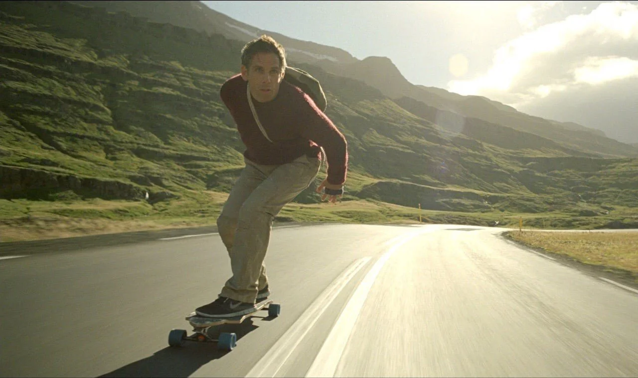 Un homme faisant du skateboard sur une route de campagne entourée de montagnes avec un ciel en partie nuageux.