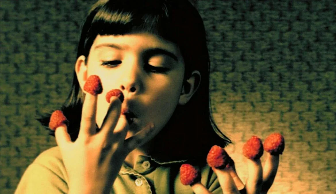 Une jeune femme avec des cheveux courts noirs mangeant des fraises, avec plusieurs fraises également posées sur ses doigts.