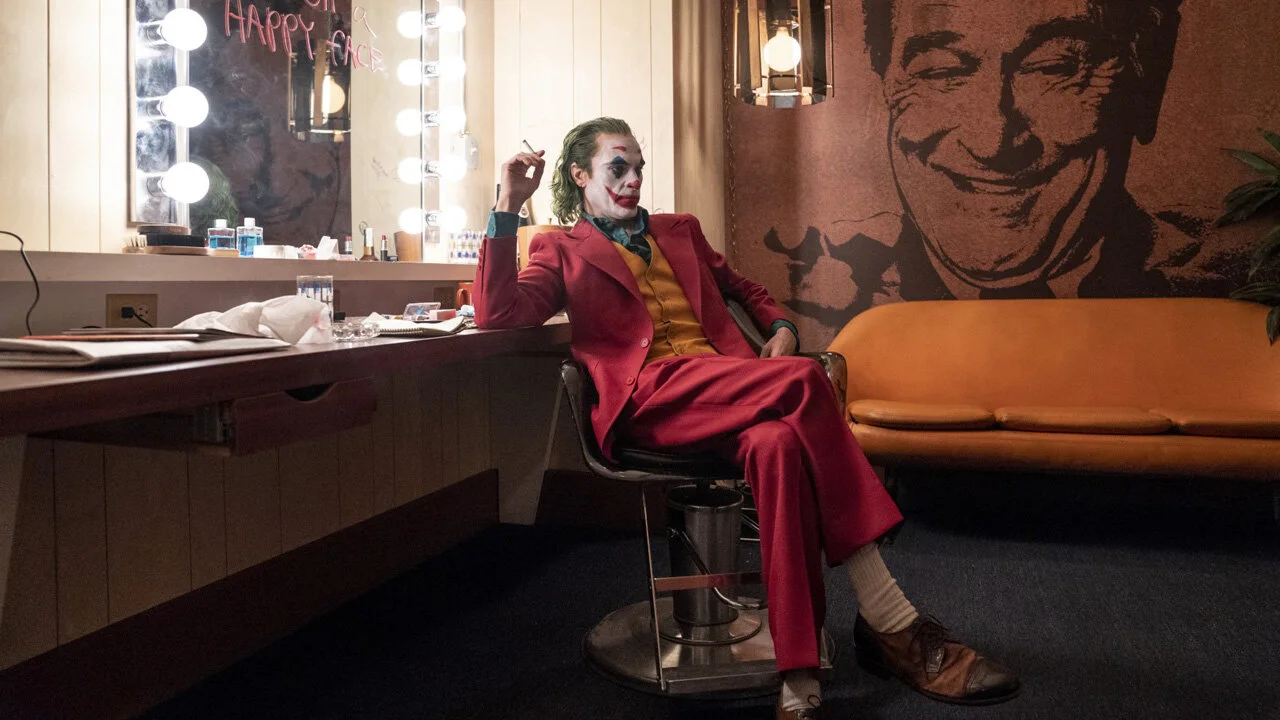 Personnage déguisé en Joker assis dans une loge de maquillage avec un maquillage de joker, portant un costume rouge, dans un décor avec un mur en mosaïque de photo d'un homme souriant.