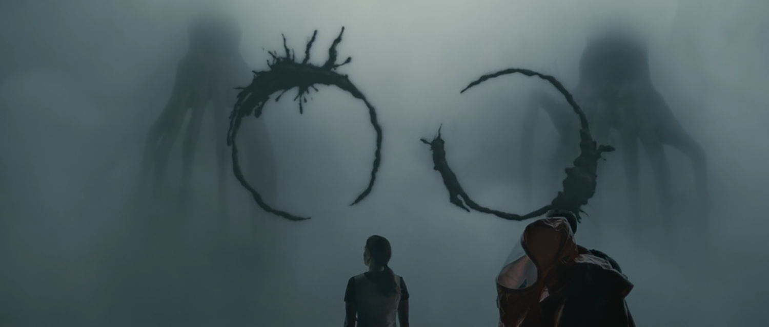 Une personne debout devant une grande affiche de film représentant deux créatures monstrueuses avec le logo du film. La personne porte une capuche, et l'atmosphère est sombre et mystérieuse.