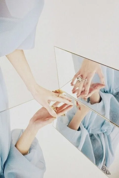 Deux mains manipulent des bijoux devant un miroir, réflexion montrant la personne portant une robe bleue et un bague.