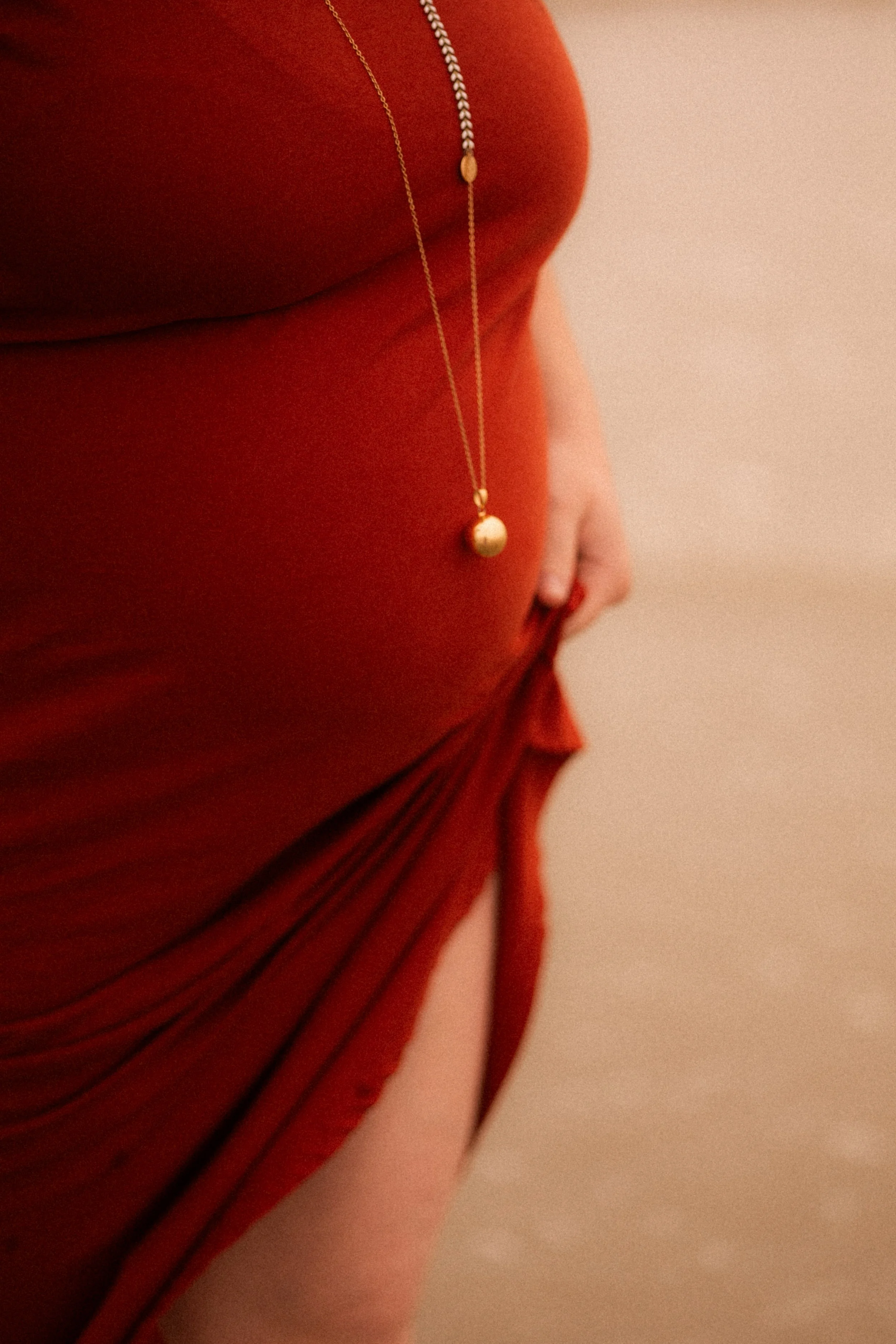 Partie du corps d'une personne portant une robe rouge, avec un collier doré et la main posée sur la hanche.