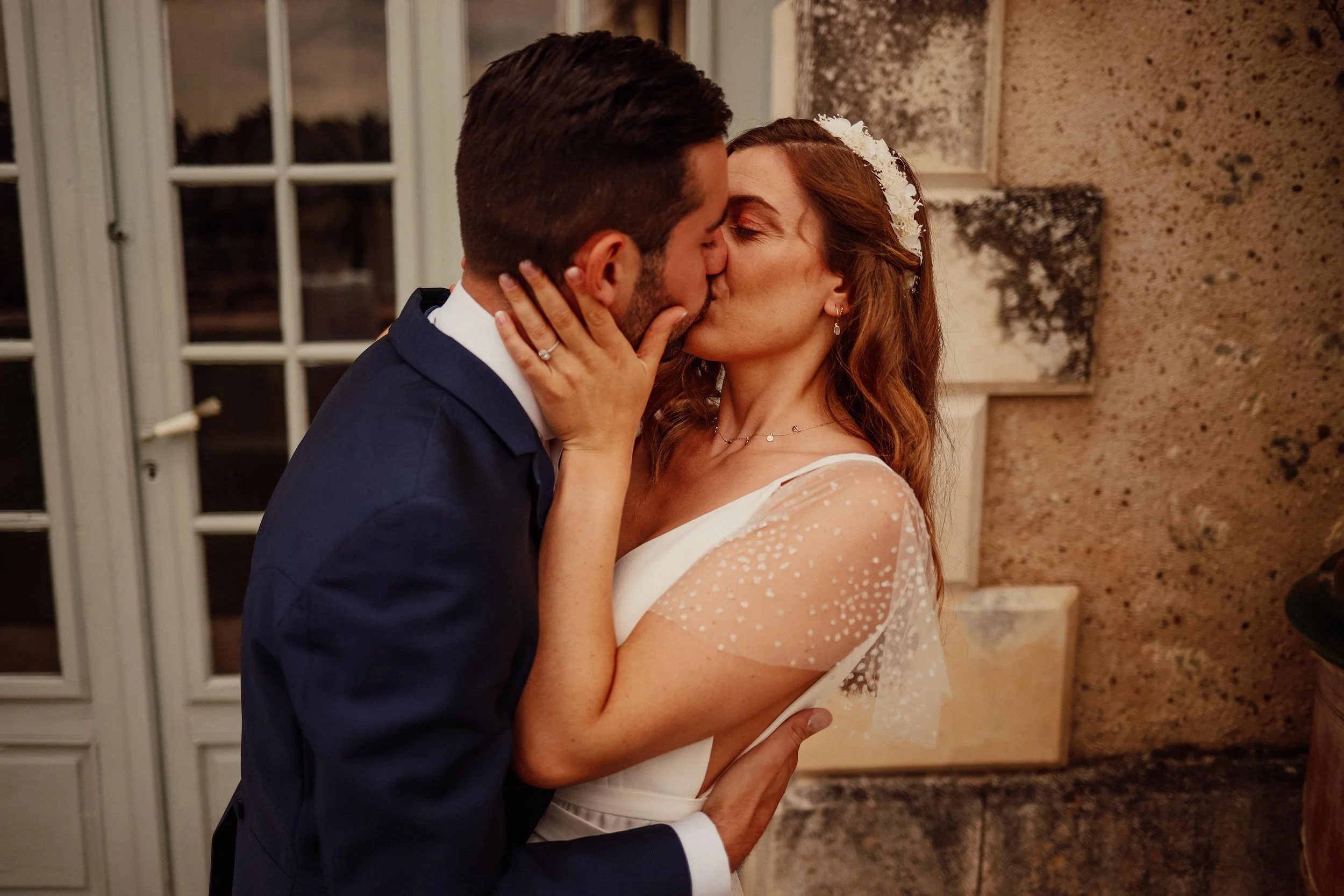 Un couple en costume de mariage s'embrasse lors de leur mariage, la femme porte une robe blanche avec des manches en tulle et un voile, l'homme porte un costume bleu foncé.